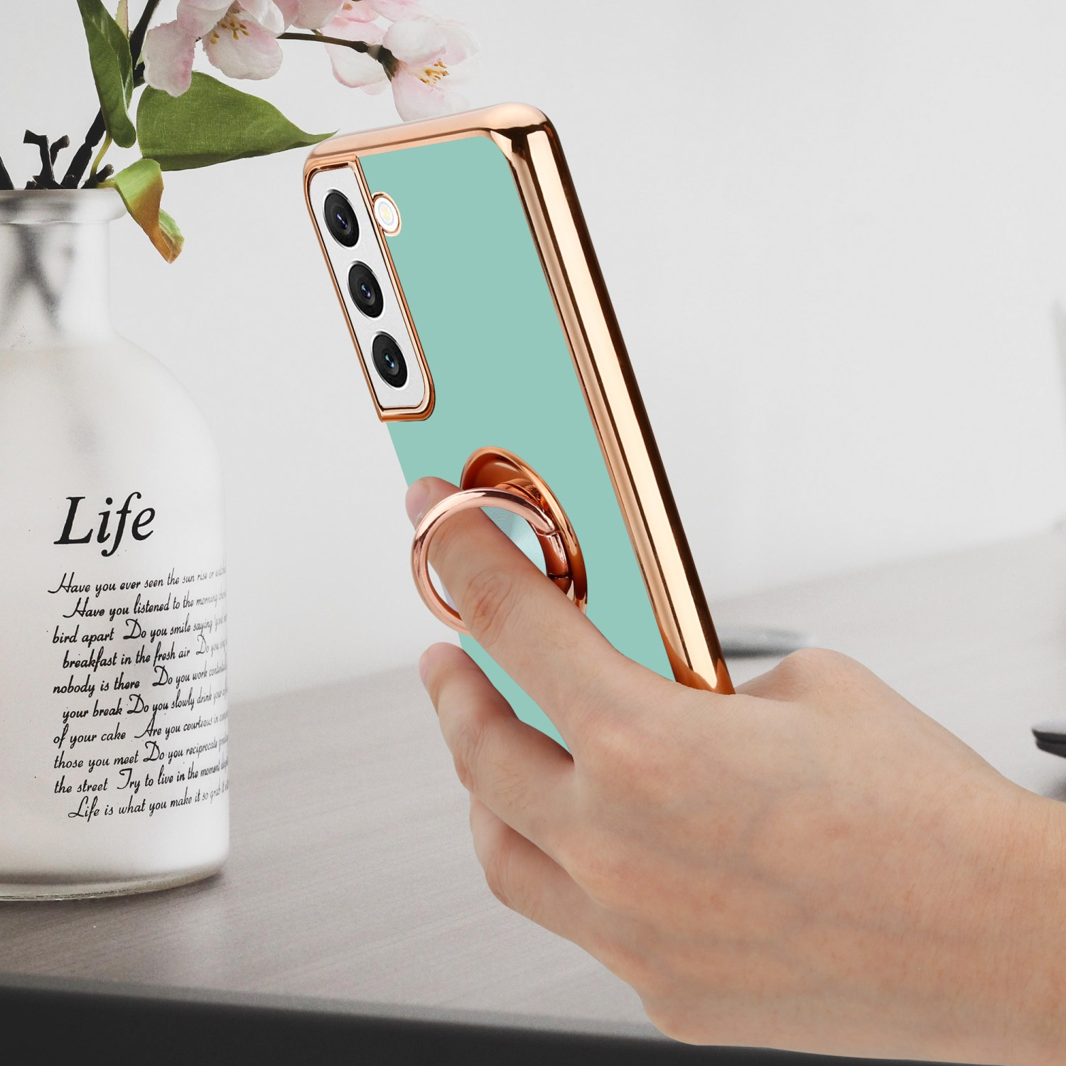 Una mano che tiene un telefono verde menta con un anello d'oro. Un vaso bianco e fiori sullo sfondo.