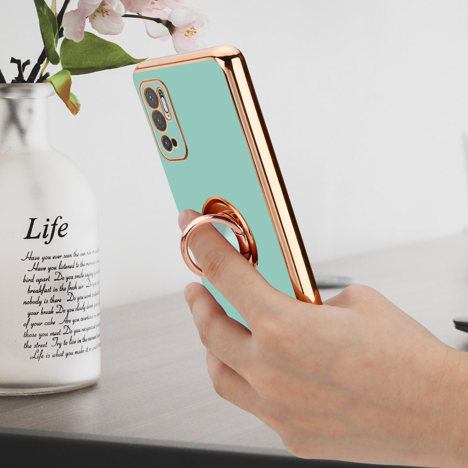 Mano che tiene una custodia per telefono con bordo dorato e anello. Sfondo con vaso e fiori.
