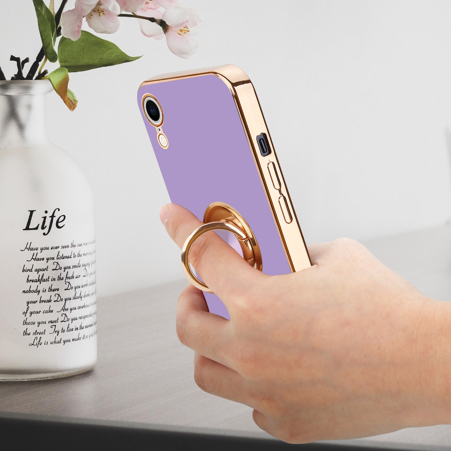 Hand hält ein lila Telefon mit goldenem Ringhalter, Blumenvase im Hintergrund.