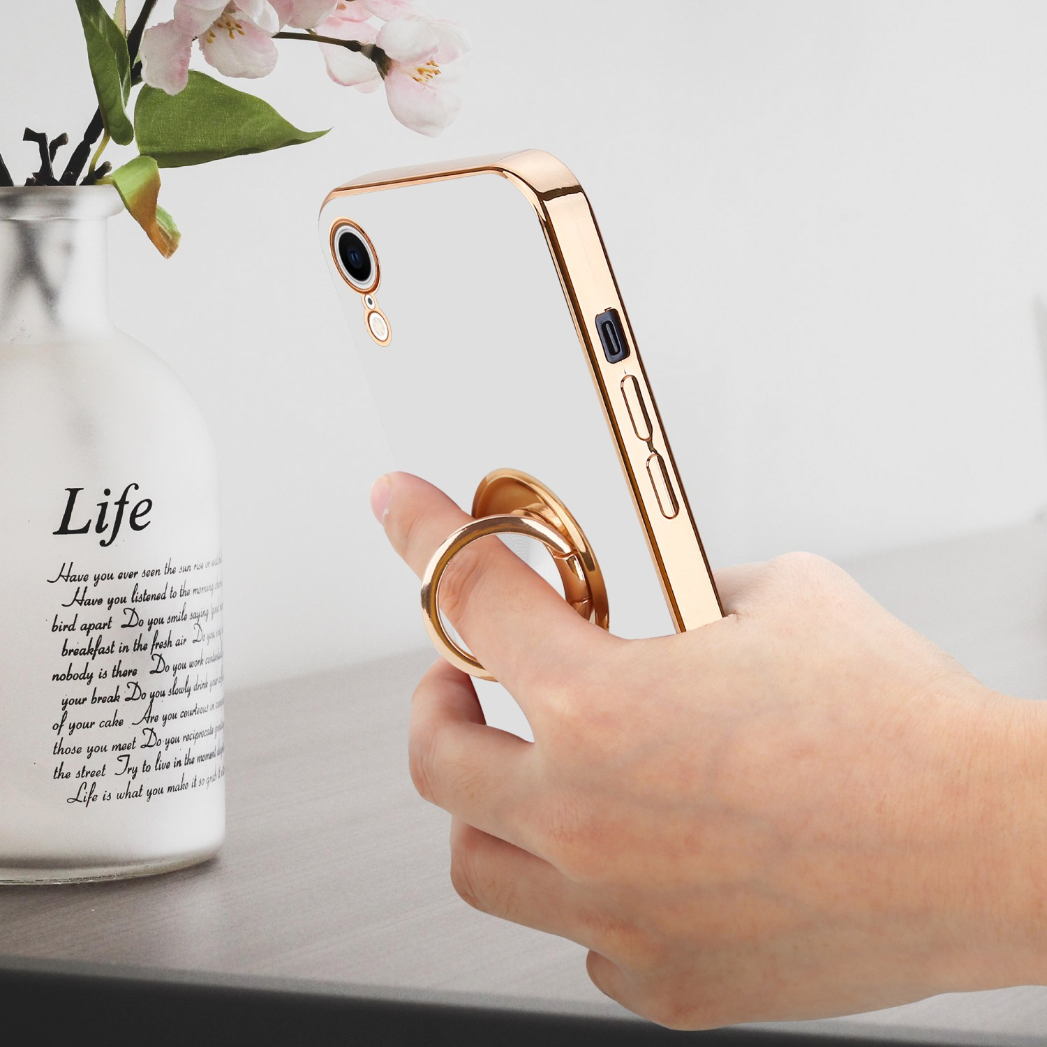 Eine Hand hält ein Telefon mit weißer Hülle und goldenem Rahmen. Links eine Vase mit Blumen.