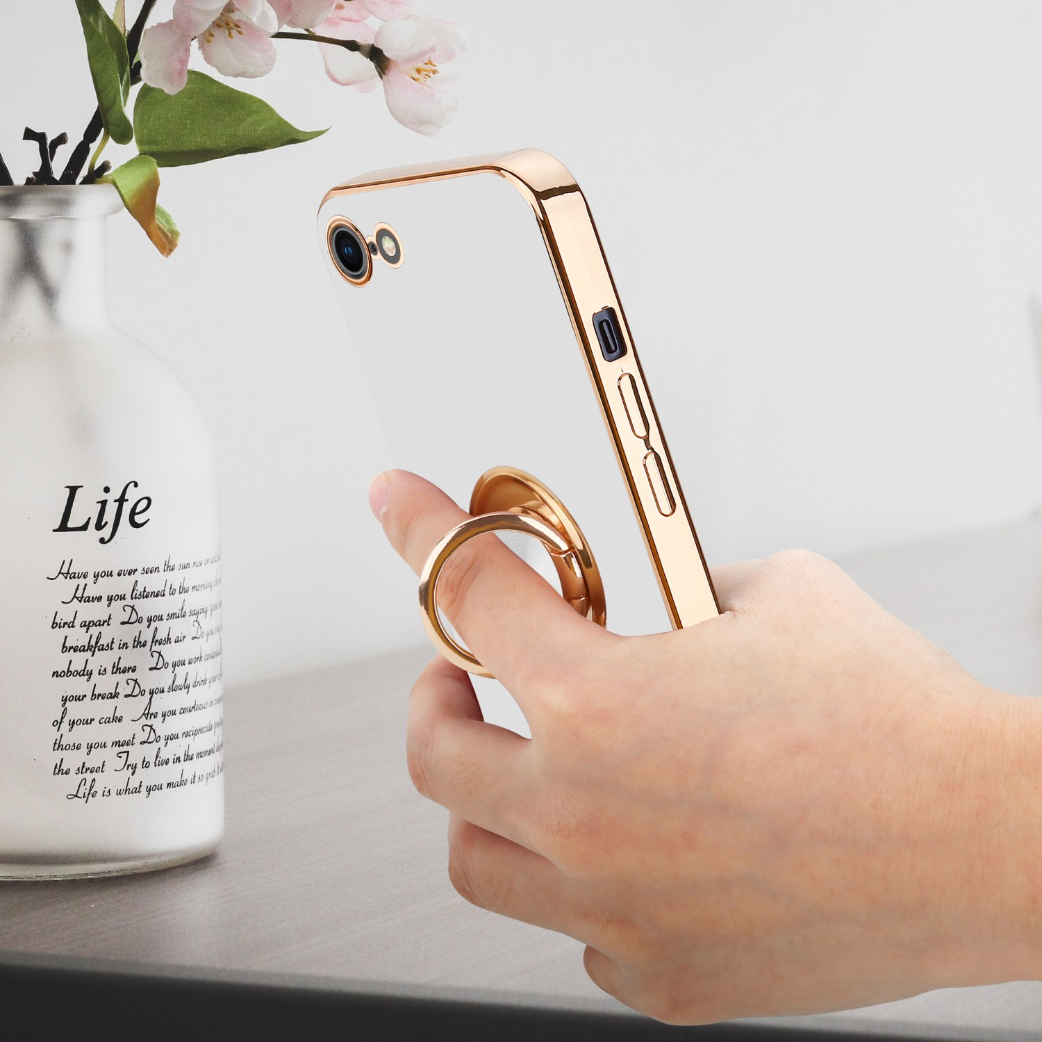 Eine Hand hält eine goldgerahmte Handyhülle. Das Telefon steht auf einem Tisch mit einer Vase und Text.