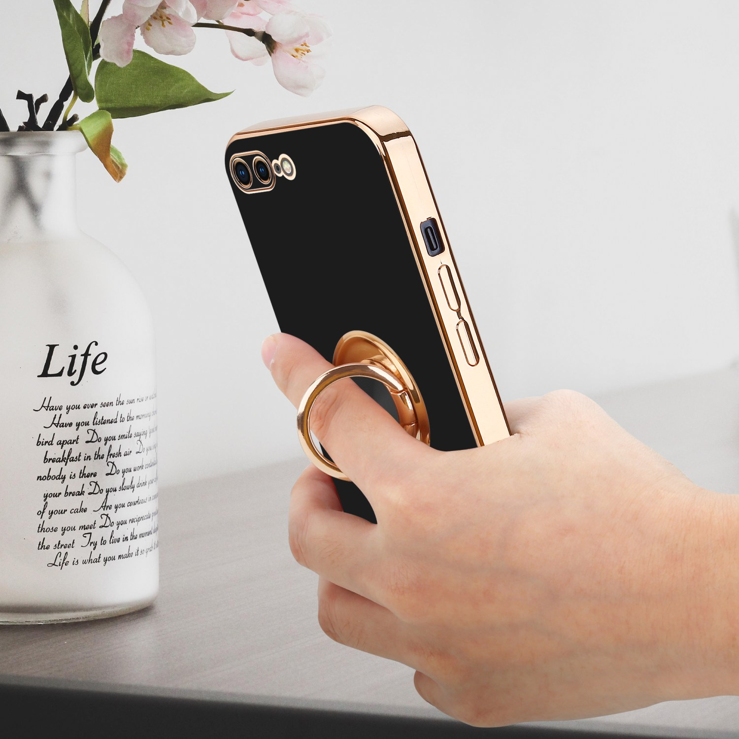 Eine Hand hält ein Telefon mit goldenem Ringhalter. Im Hintergrund eine Vase mit Schrift.