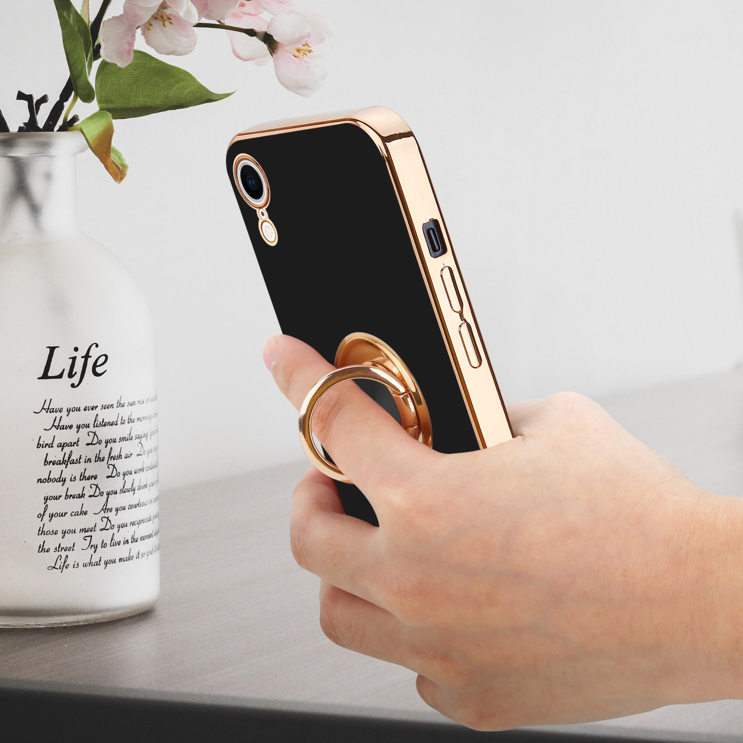 Hand, die ein Telefon mit einem Ringhalter hält. Es gibt eine Vase und einige Wörter darauf.