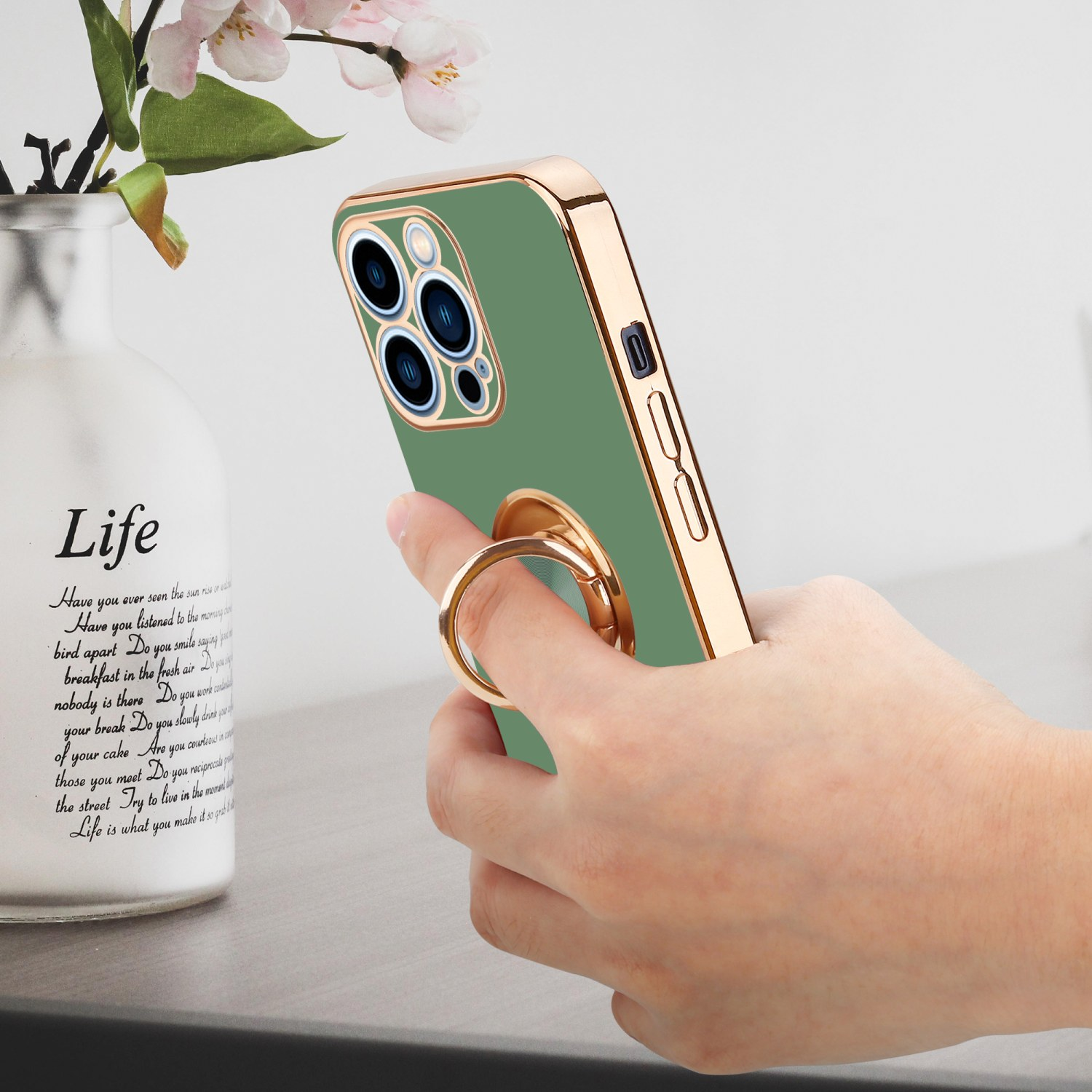 Eine Hand, die ein grünes Telefon mit goldenem Ringständer hält, neben einer Vase.