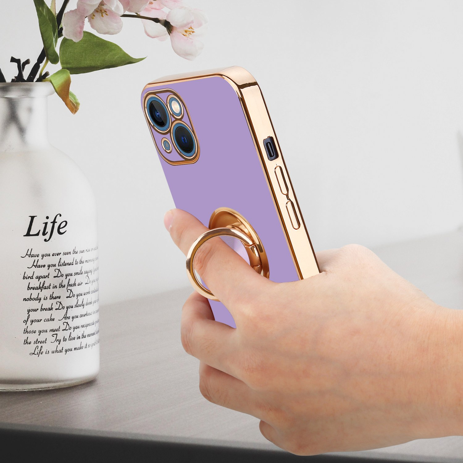 Hand, die ein Telefon mit lila Hülle und goldenem Ringhalter hält, im Hintergrund eine Vase mit Blumen.
