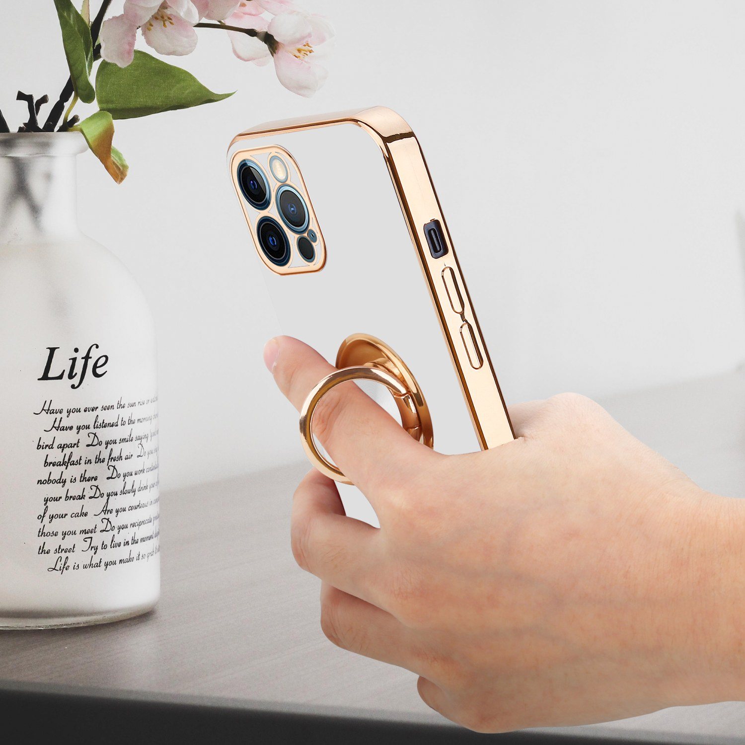 Eine Hand hält ein goldgerahmtes Telefon. Links steht eine weiße Vase mit Blumen.