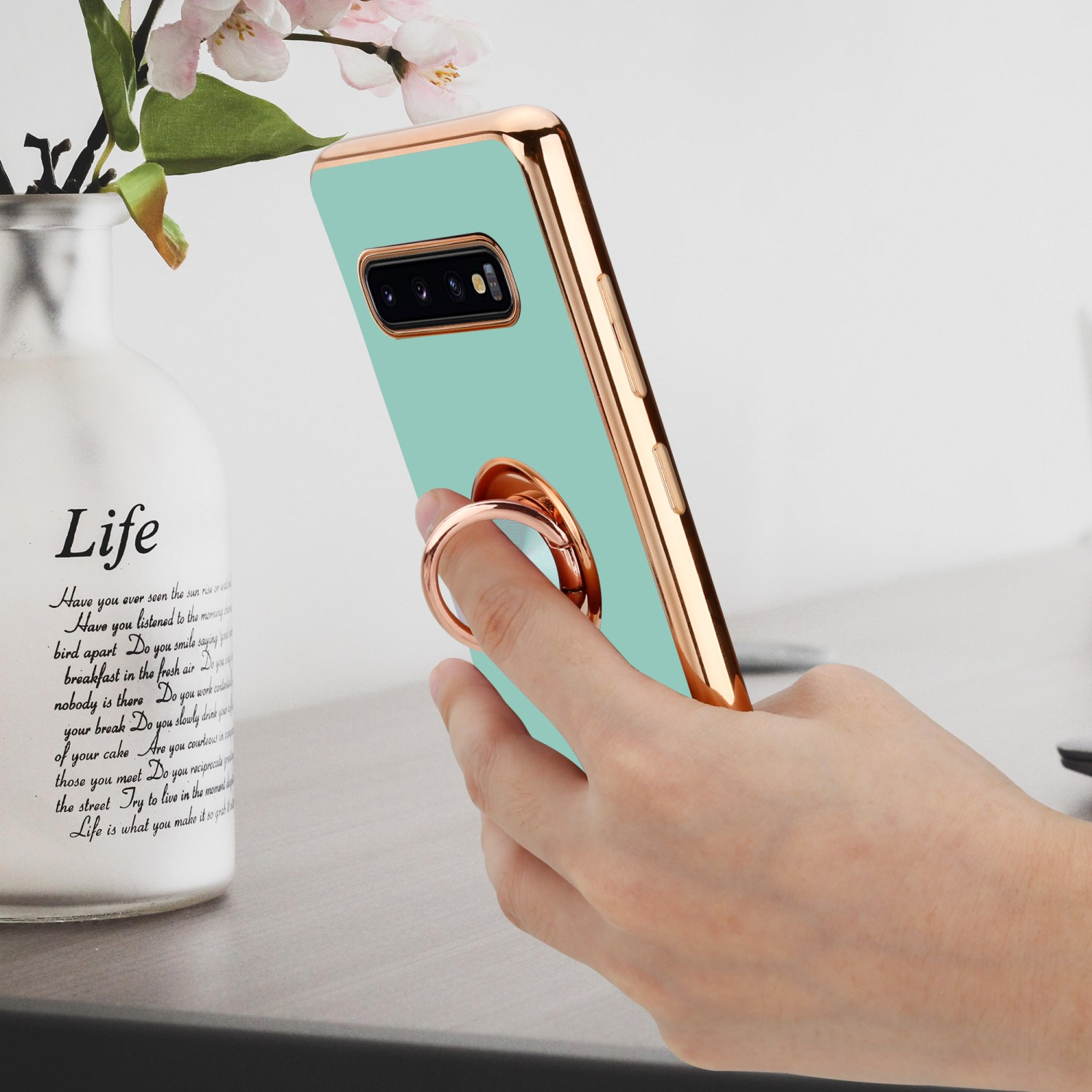 Una mano che tiene un telefono verde menta con un'impugnatura ad anello dorata, accanto a un vaso bianco.