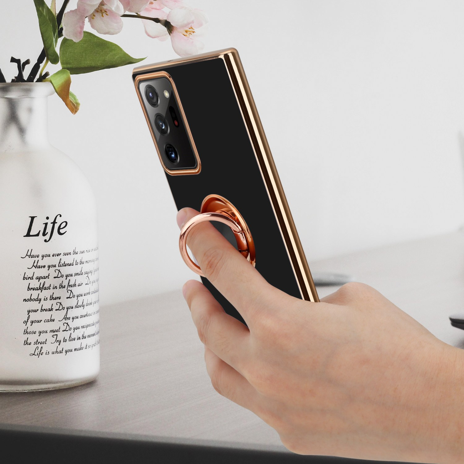 Eine Person hält ein schwarzes Telefon mit goldenem Ringhalter. Eine Vase mit Blumen ist im Hintergrund.