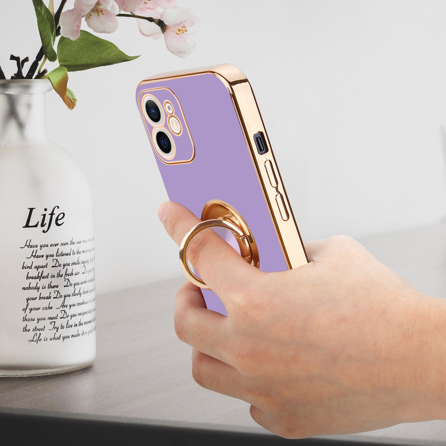 Eine Hand, die ein lila Telefon mit goldenem Ringhalter hält, mit Vase im Hintergrund.