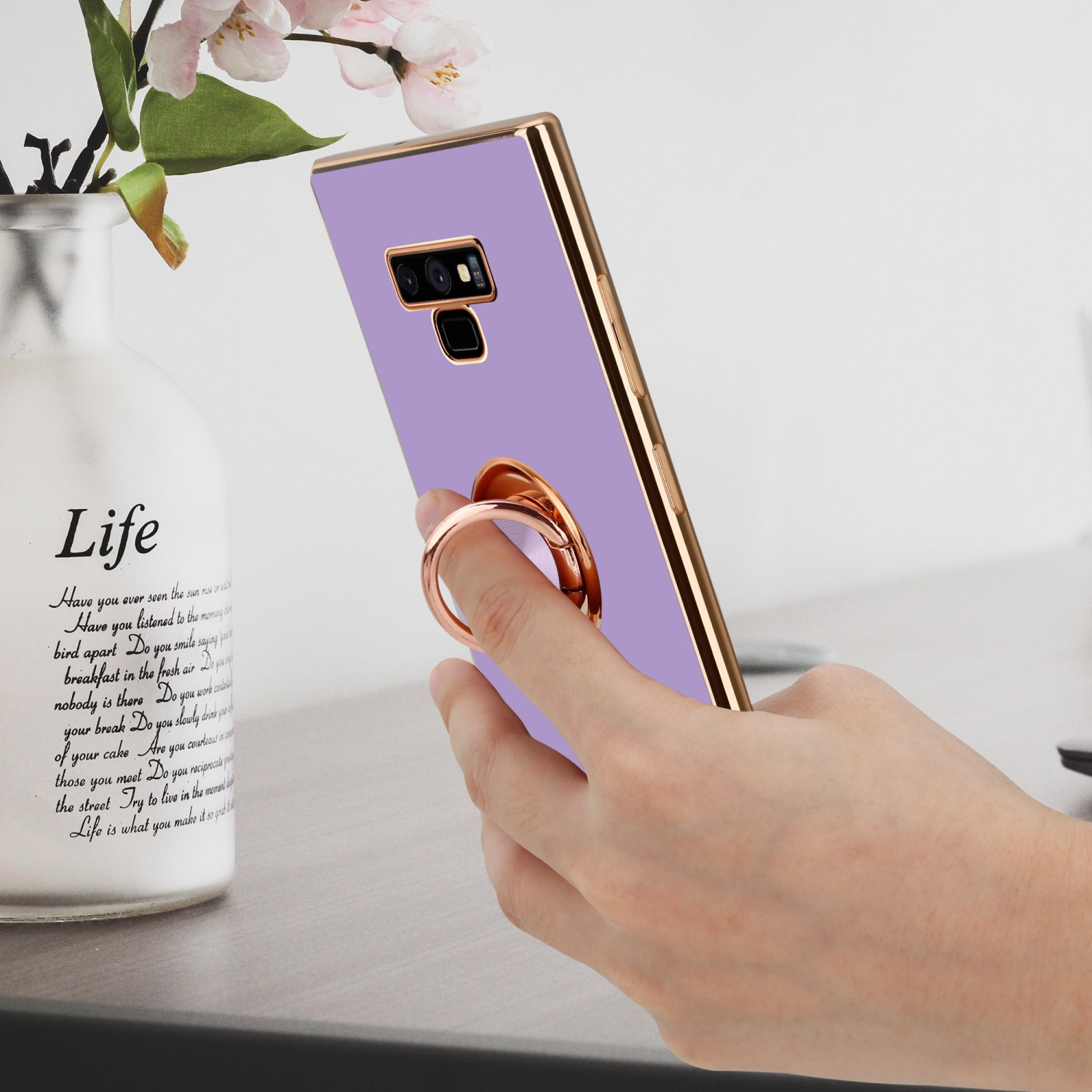Una mano che tiene un telefono con custodia viola e cornice dorata. A sinistra, un vaso con fiori.