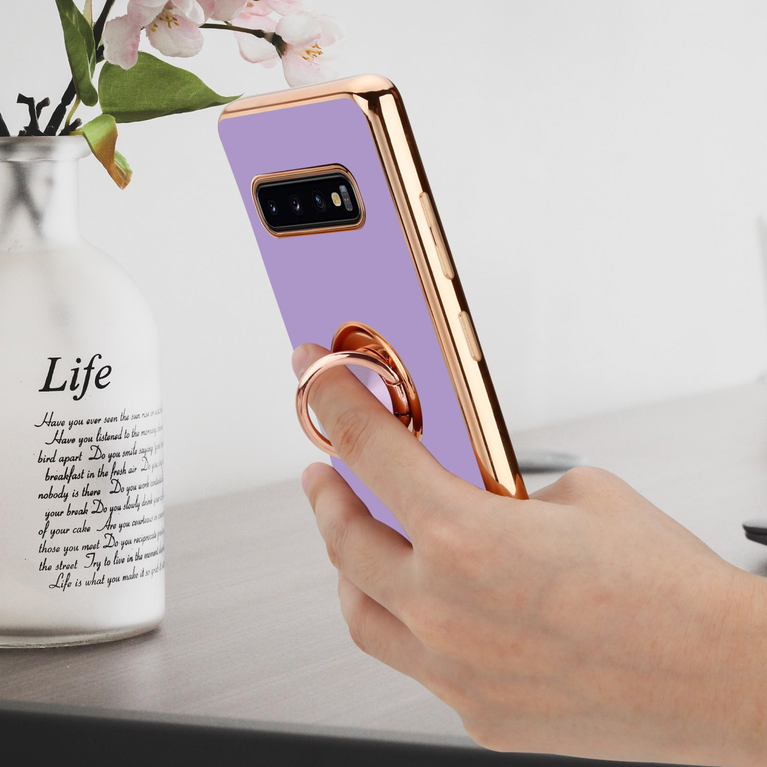 Hand, die lila Telefon mit Ringhalter hält. Weiße Vase und Blumen auf dem Tisch.