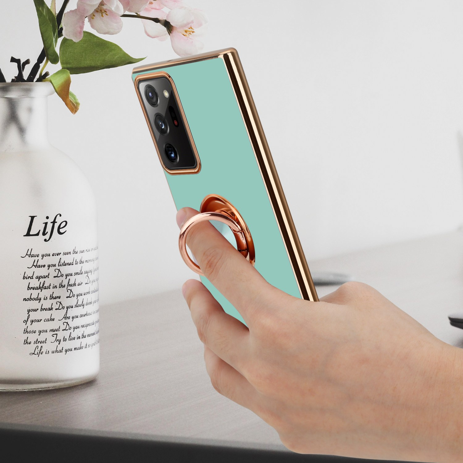 Eine Hand hält eine Handyhülle mit Ring. Die Hülle ist mintgrün und goldfarben. Eine Vase steht auf dem Tisch.