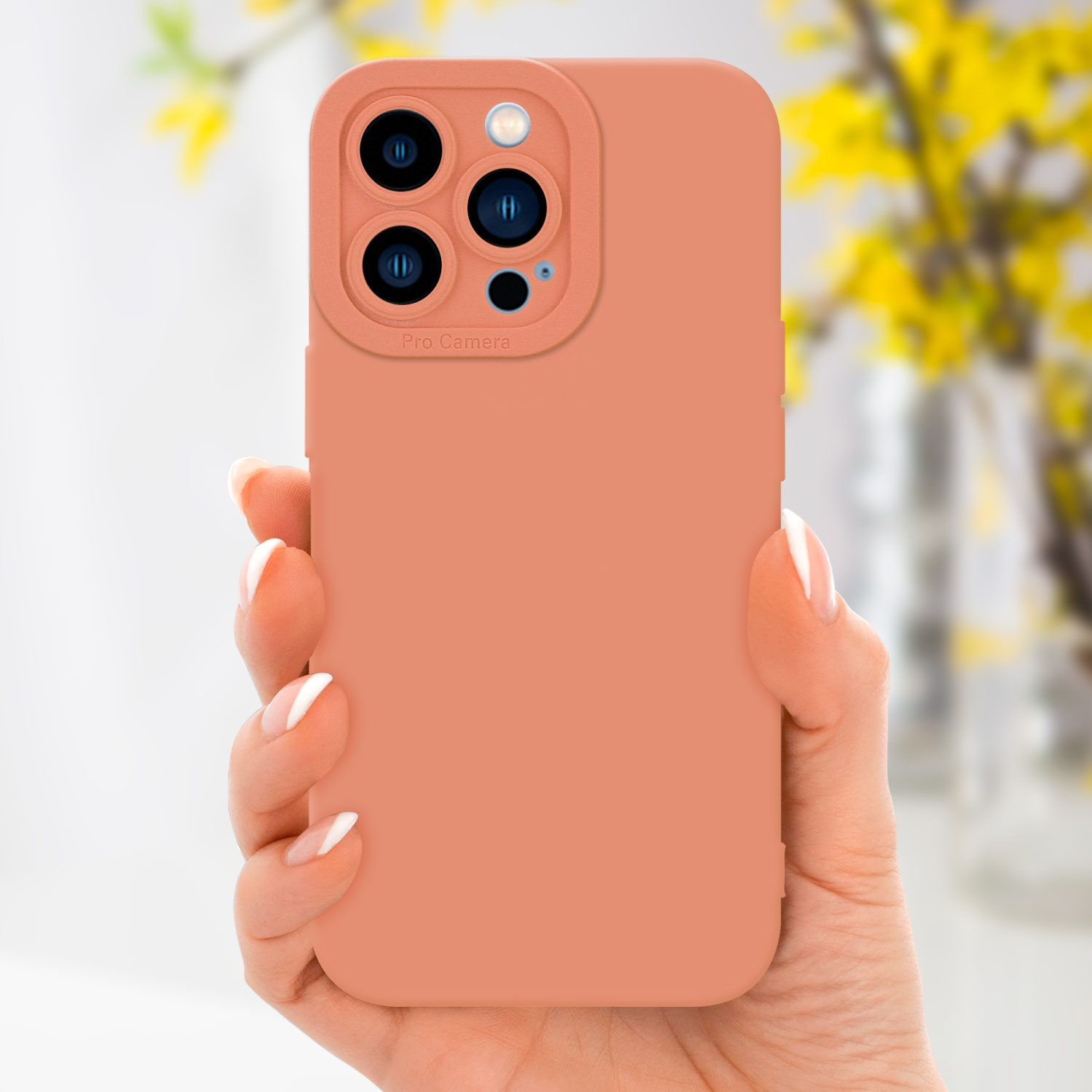 Una mano tiene una custodia per telefono color pesca con dettagli della fotocamera. Lo sfondo ha fiori sfocati.