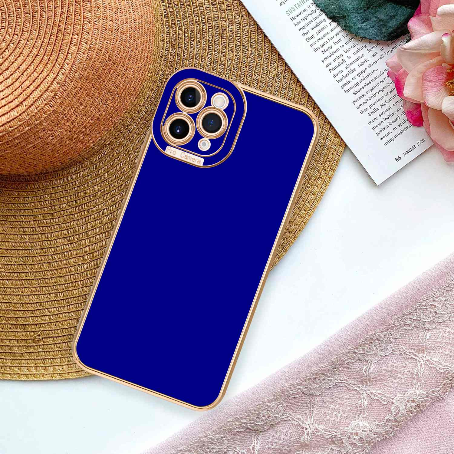 Blaue Handyhülle mit goldfarbenem Rand und Strohhut auf einem Tisch mit Buch und rosa Blume.