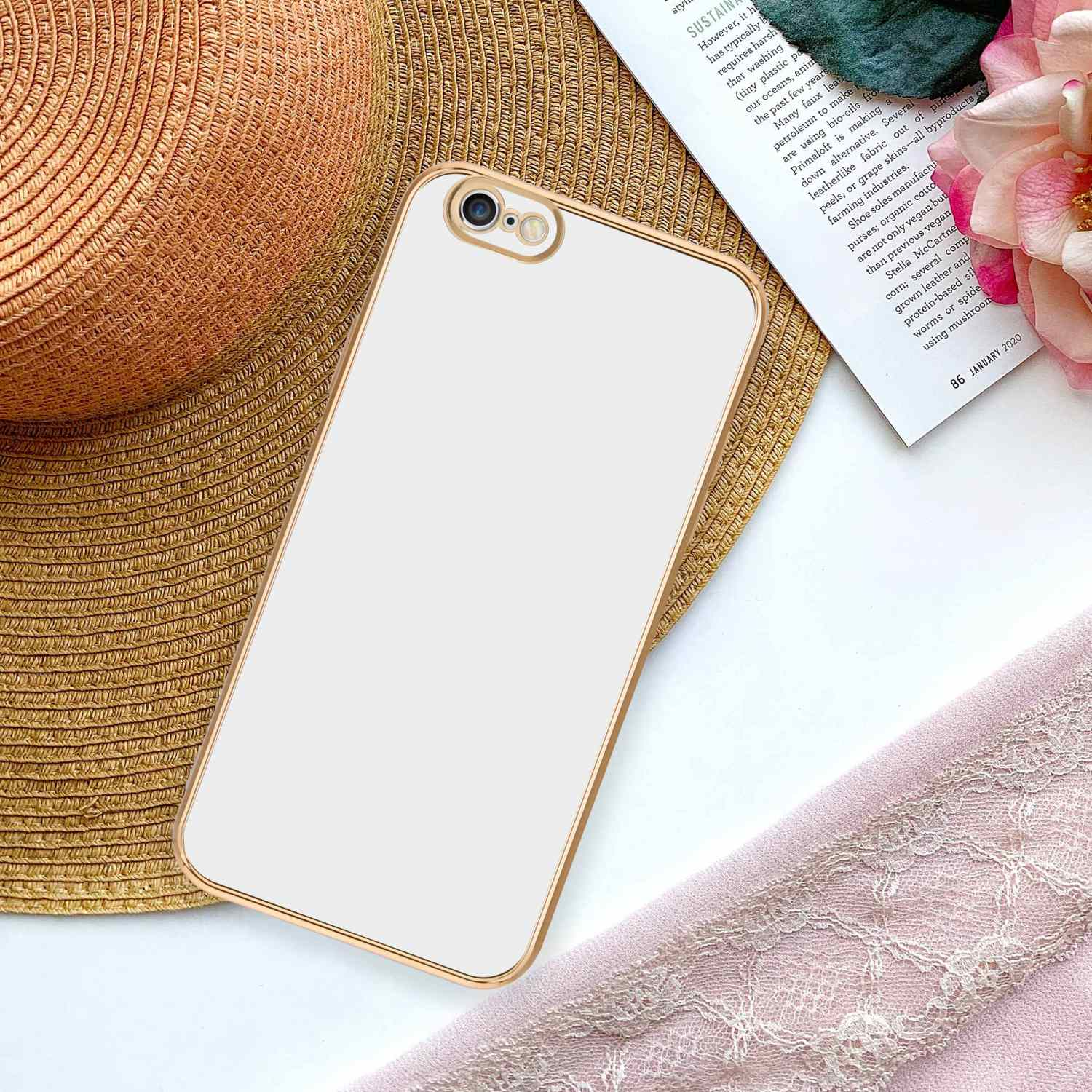 Telefon in goldgerahmter weißer Hülle auf Tisch mit Hut, Buch und rosa Stoff.