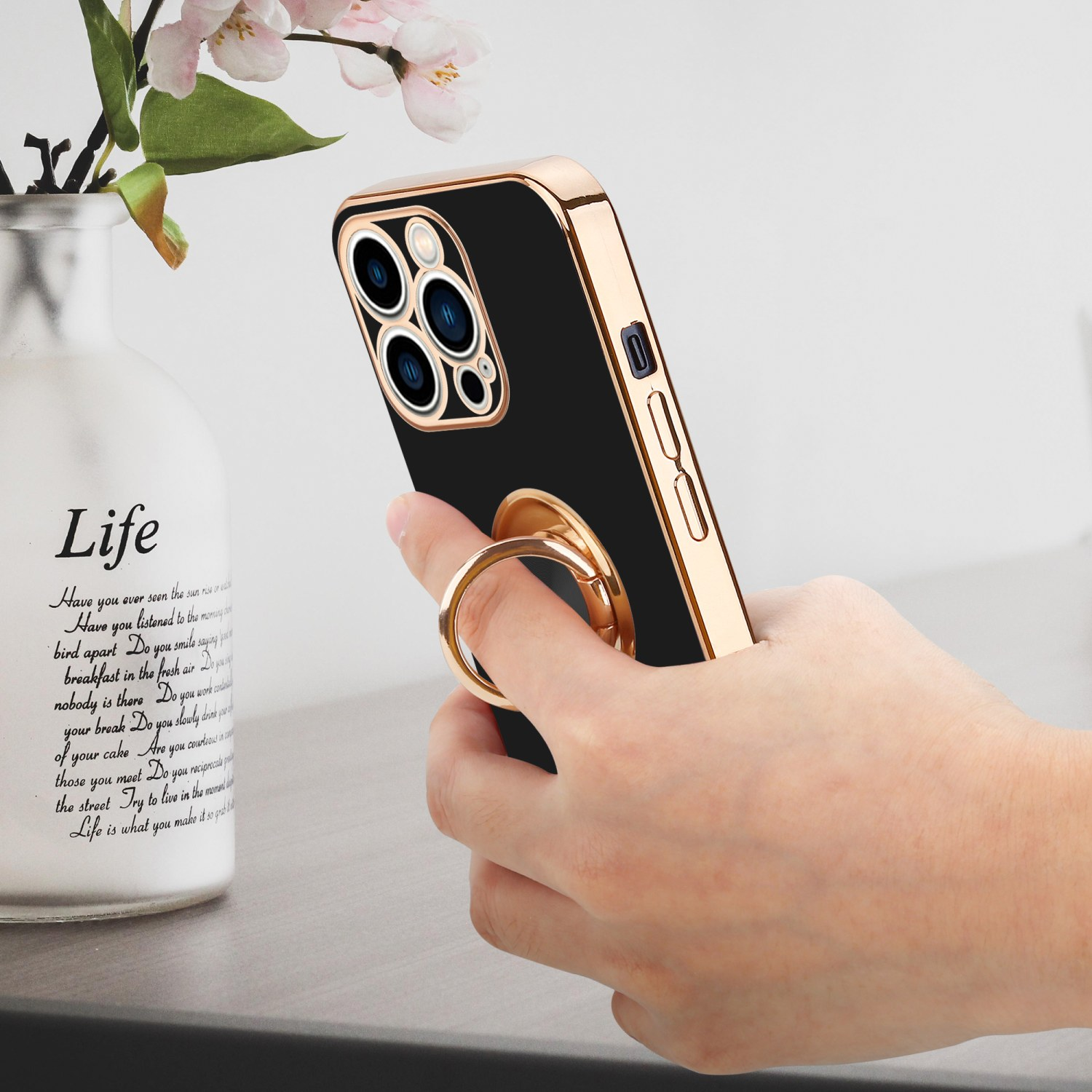 Hand hält eine schwarze Handyhülle mit goldenem Ring. Vase mit Blumen im Hintergrund.