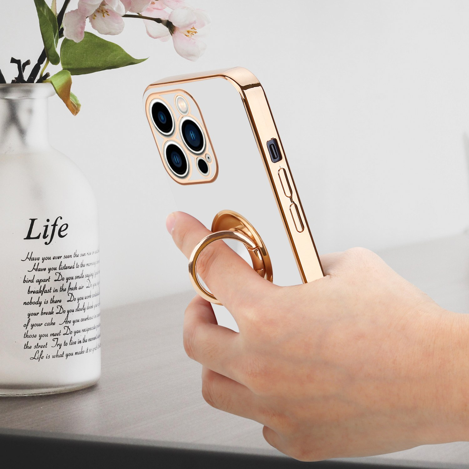 Hand hält eine goldene Handyhülle mit Ringständer neben einer Vase mit Blumen.