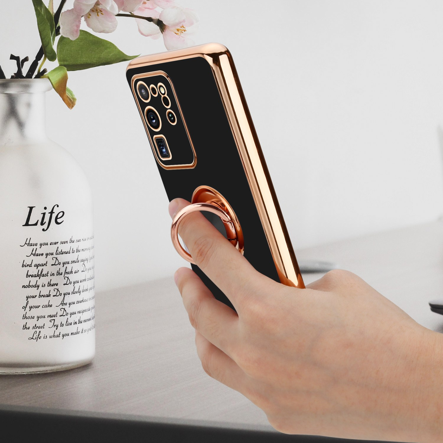 Eine Hand hält ein schwarzes Telefon mit Goldrand und Ringhalter. Es gibt eine Vase mit Blumen.
