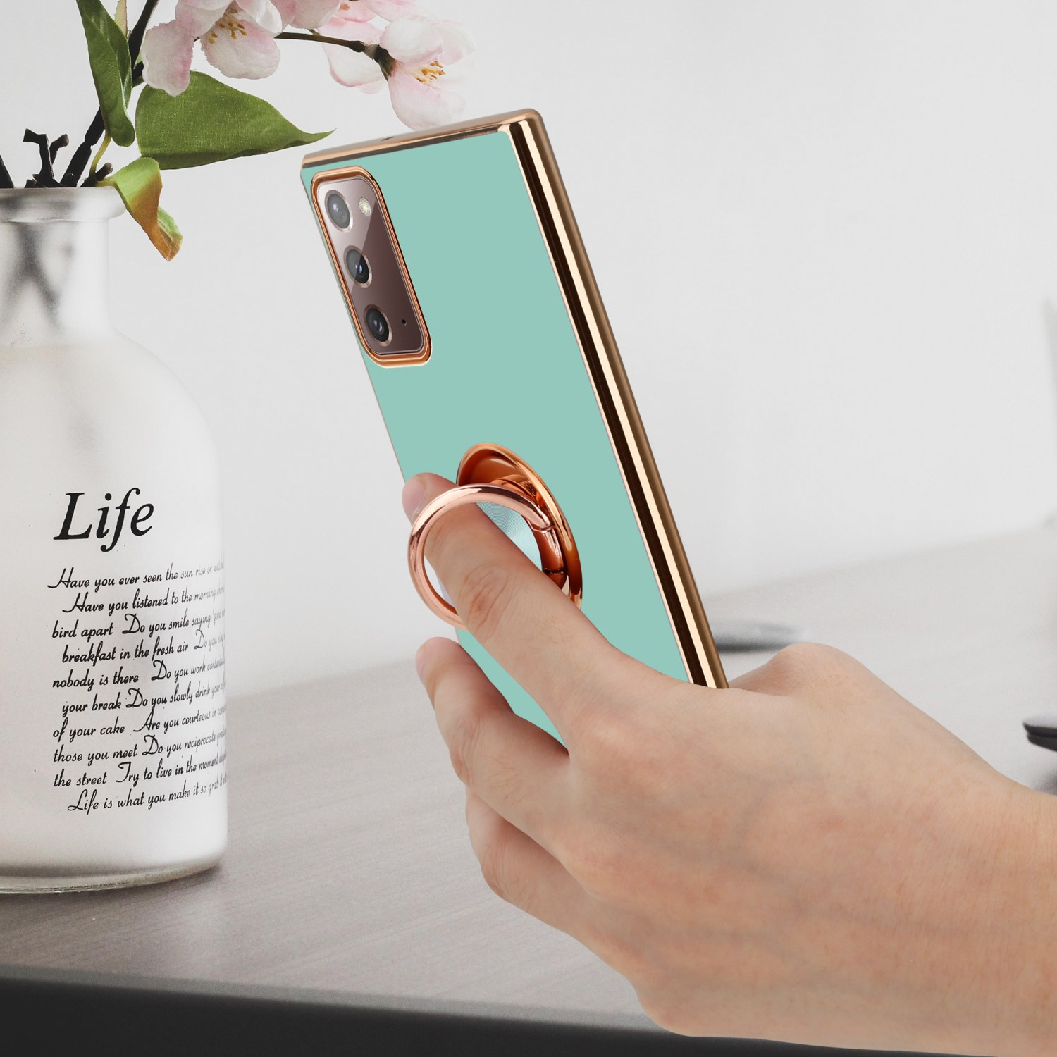 Mano di donna che tiene un telefono verde menta con un anello. Un vaso sullo sfondo.