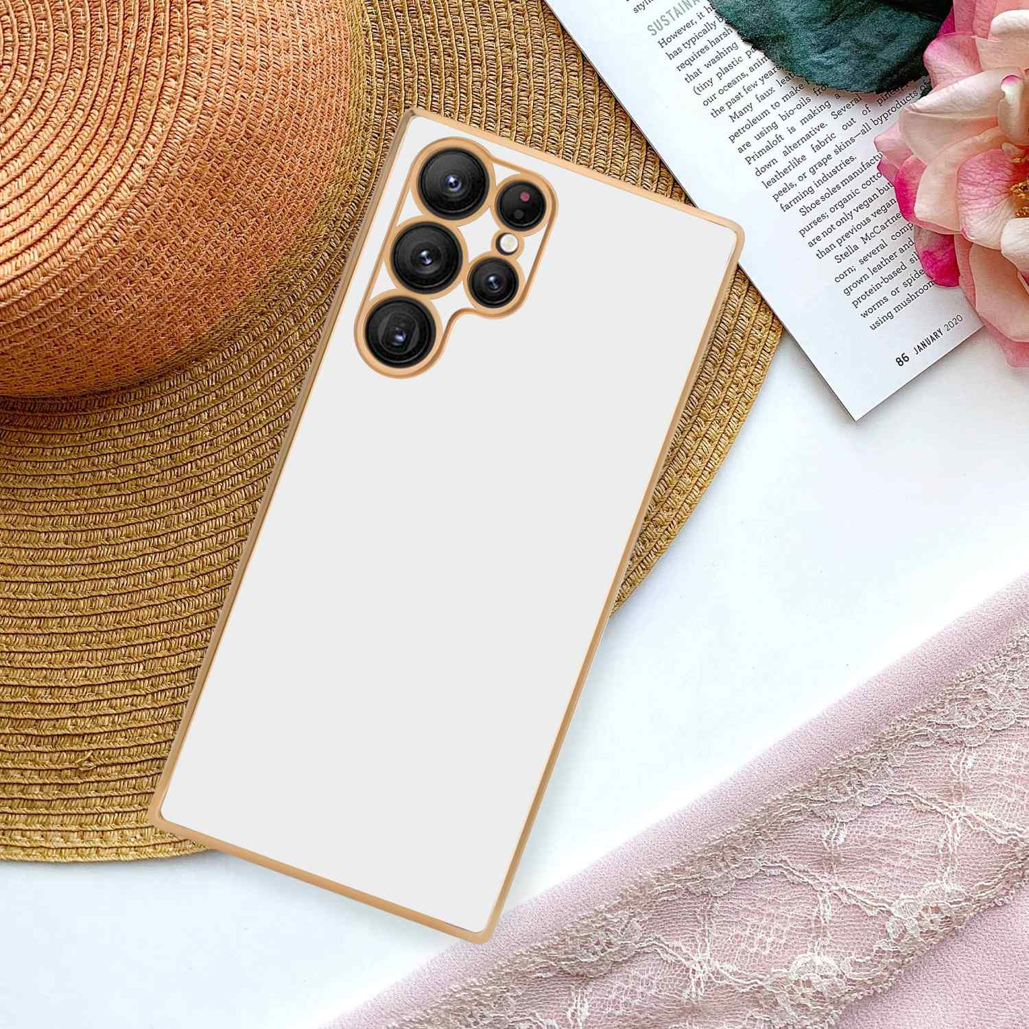 Weiße Handyhülle mit goldenem Rand. Das Telefon liegt auf einem Strohhut und einer Zeitschrift. Rosa Tuch.