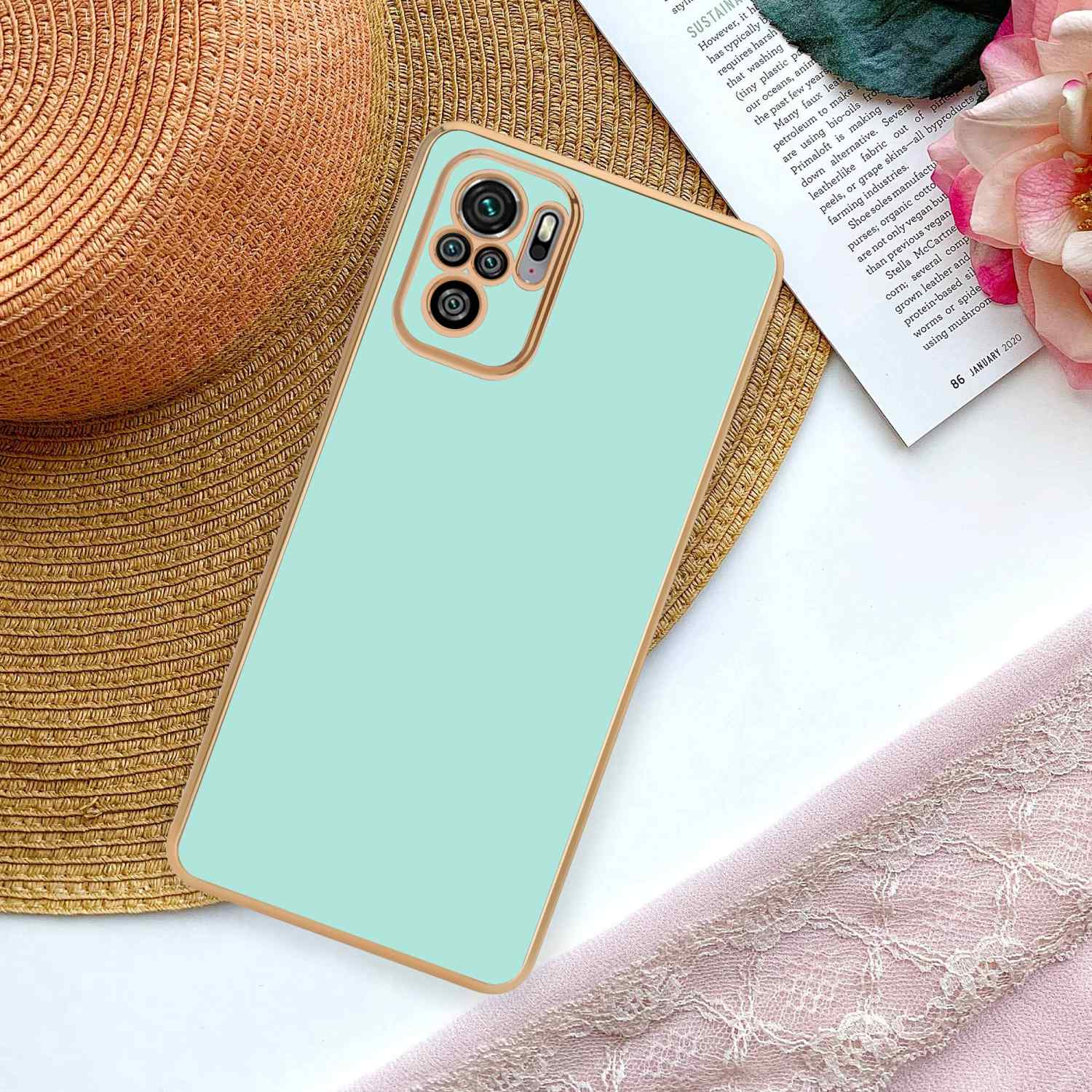 Un telefono e una custodia su una superficie con un cappello di paglia, una rivista e un tessuto rosa sullo sfondo.