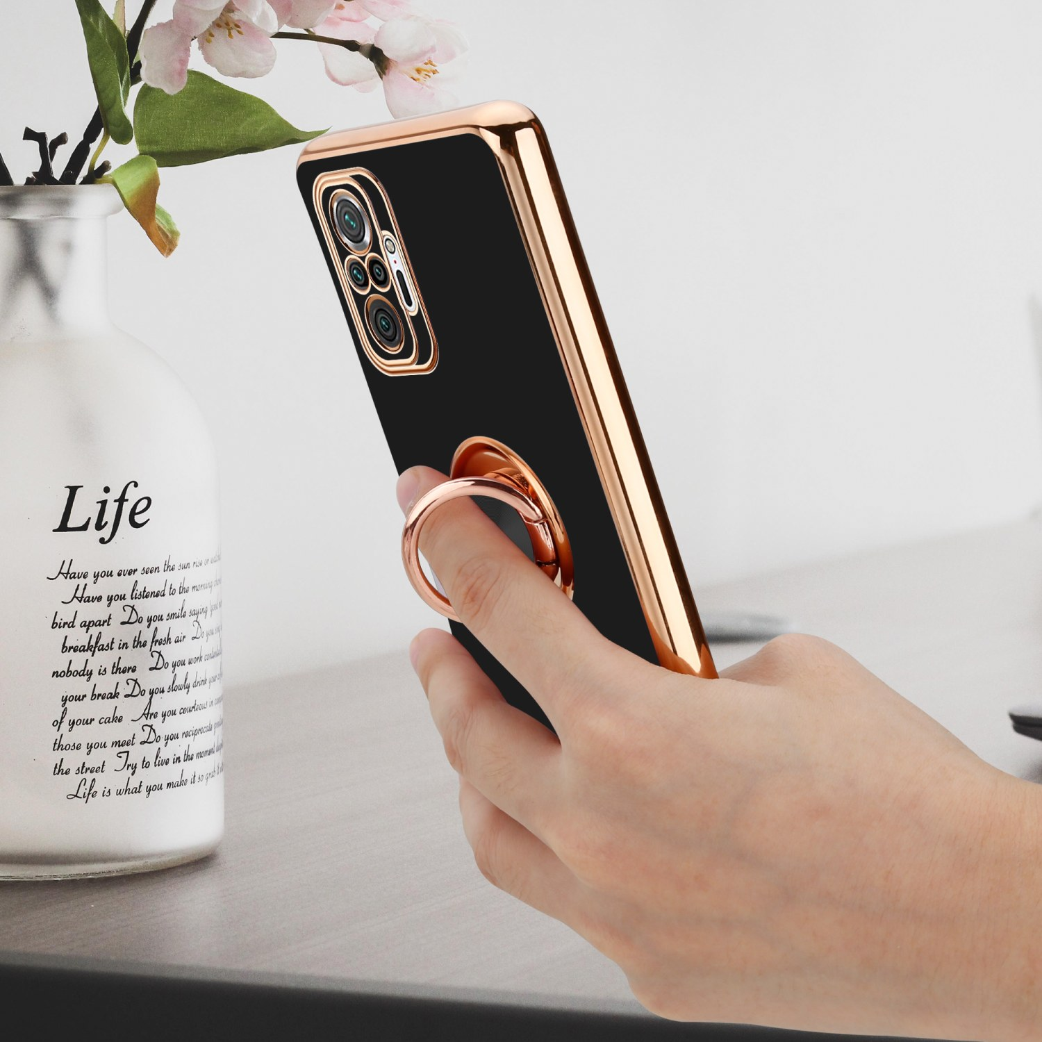 Hand hält Telefon mit schwarzer Hülle, Ringhalter, auf einem Tisch mit Vase.