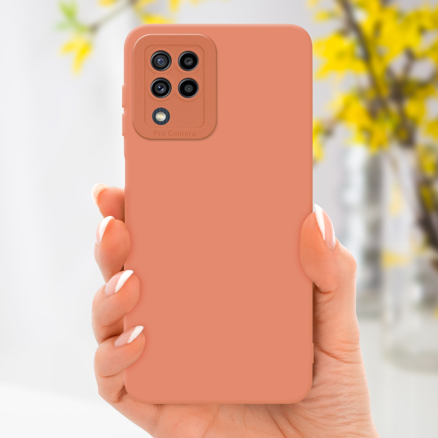 Una mano tiene una custodia per telefono color pesca con un rialzo per la fotocamera. Fiori gialli sfocati sullo sfondo.