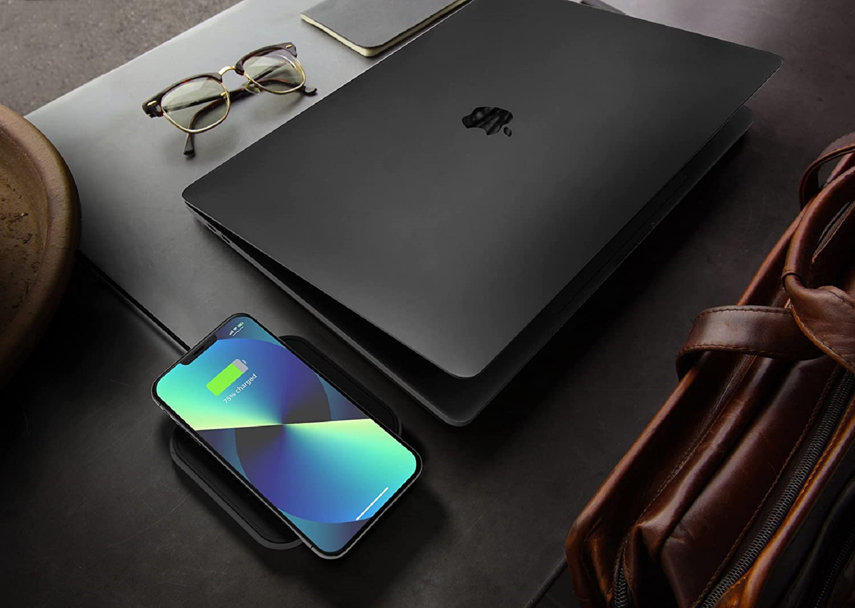 Portátil negro, teléfono en cargador inalámbrico, gafas y bolso de cuero. Portátil con logo Apple. Teléfono cargando.