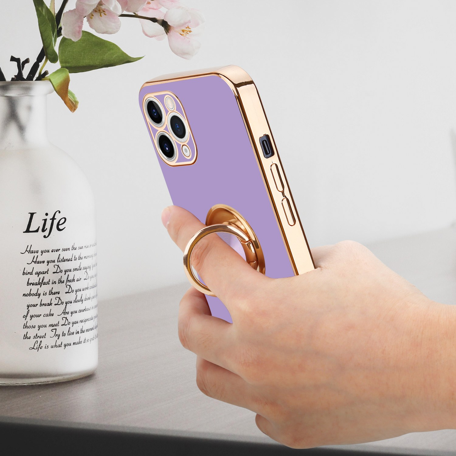Ein lila Telefon mit goldenem Ringhalter auf der Rückseite, gehalten von einer Hand, im Hintergrund eine Vase.