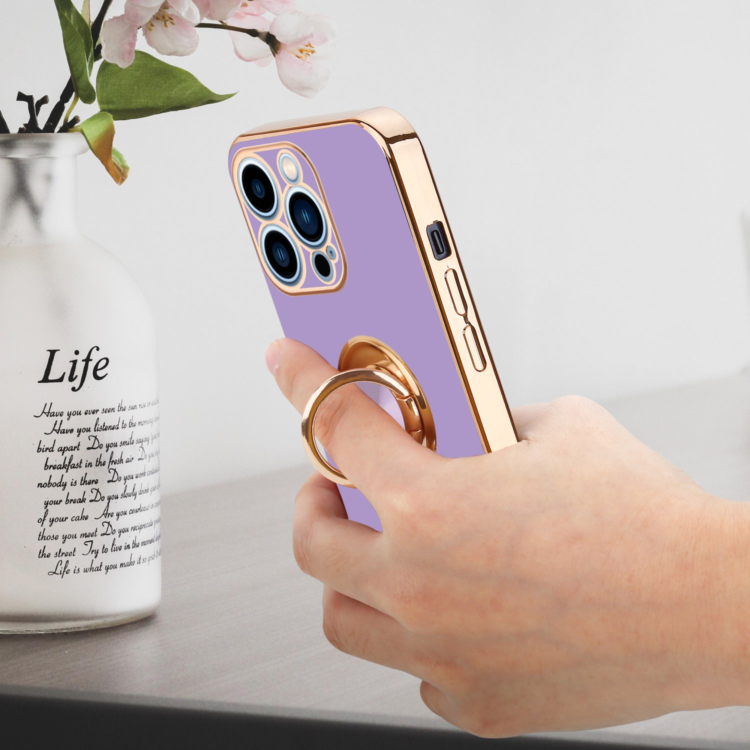 Hand hält ein lila Telefon mit goldenem Ring. Eine Vase mit Blumen ist im Hintergrund.