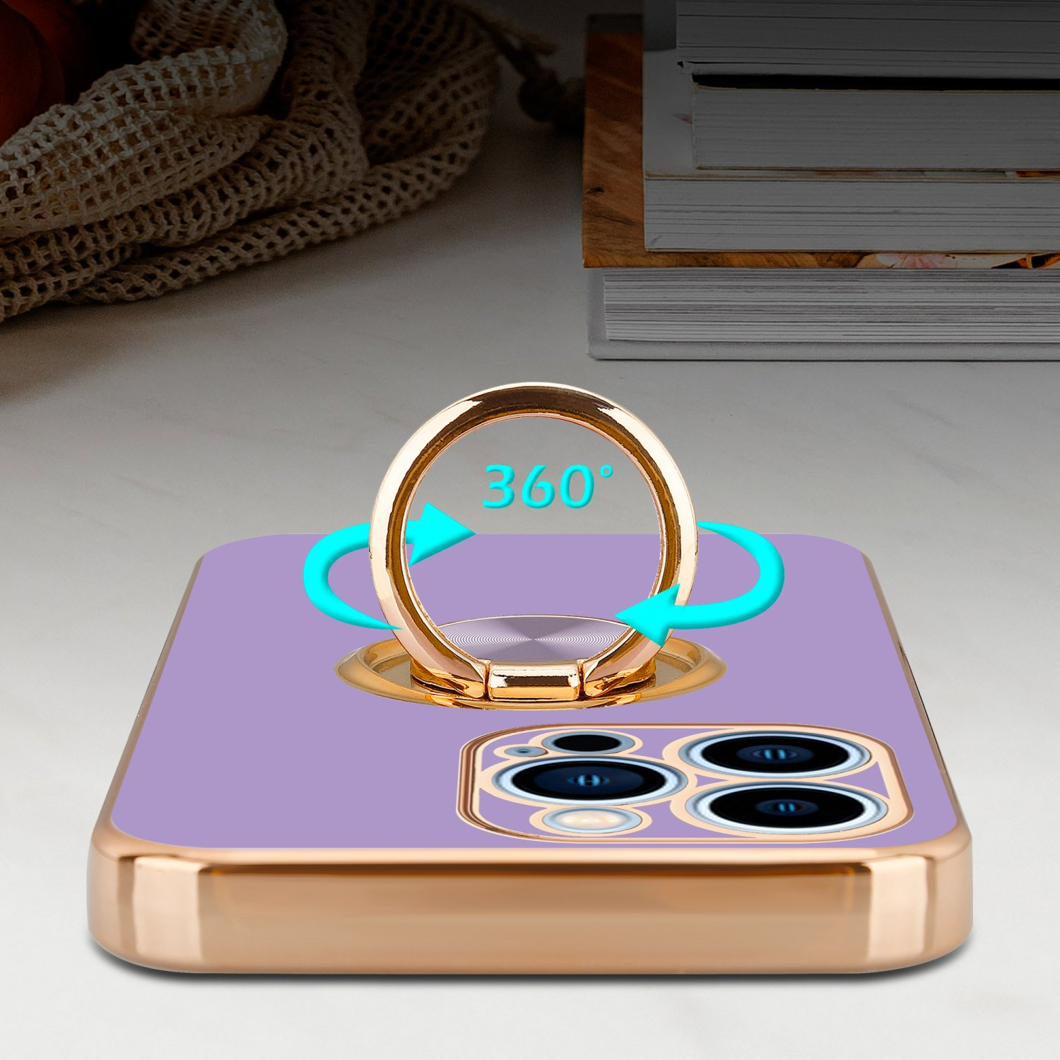 Lila Handyhülle mit goldenem Ringhalter. Hintergrund: verschwommene Bücher und Stoff.