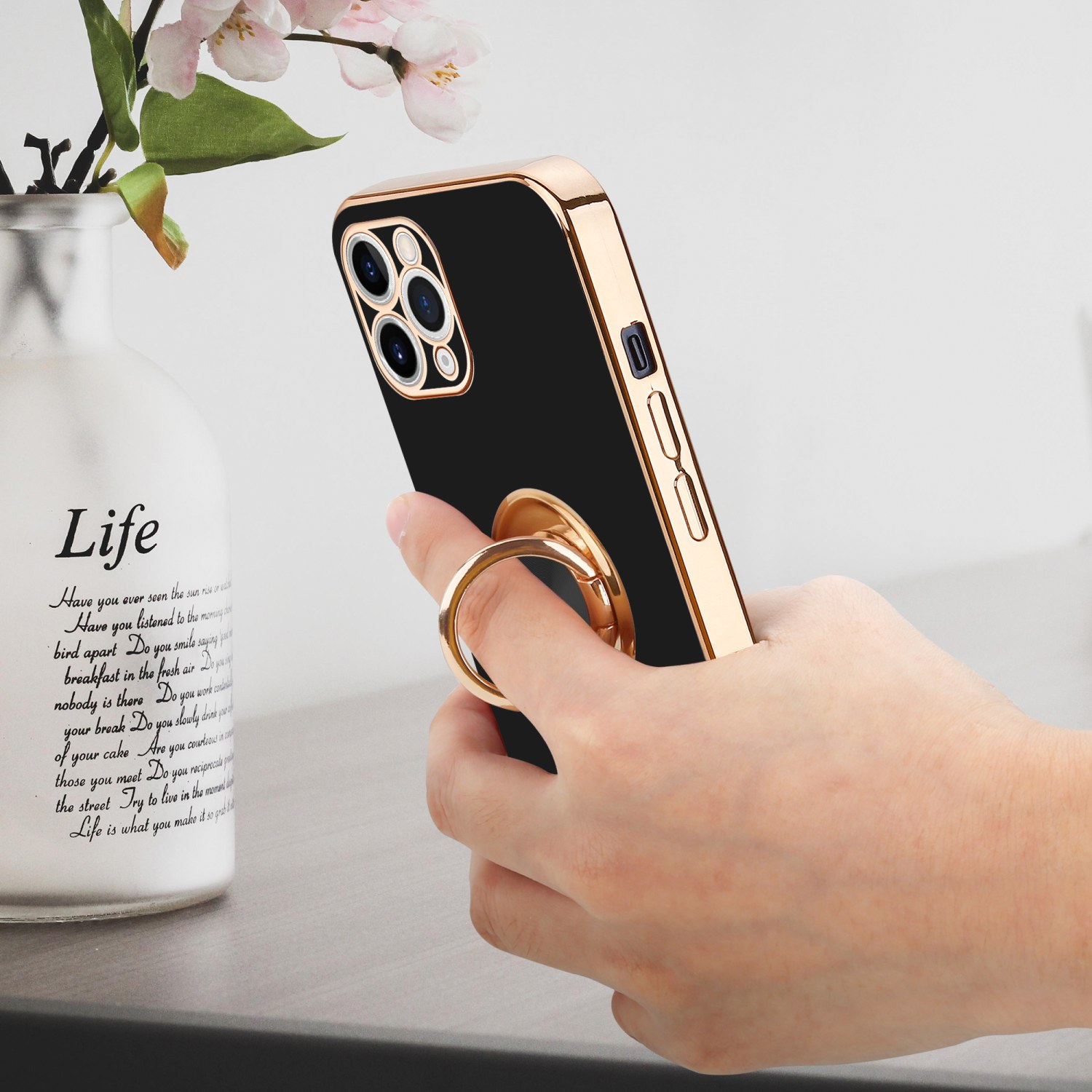 Hand hält ein schwarzes und goldenes Telefon mit Ringhalter in der Nähe einer Vase mit Blumen.