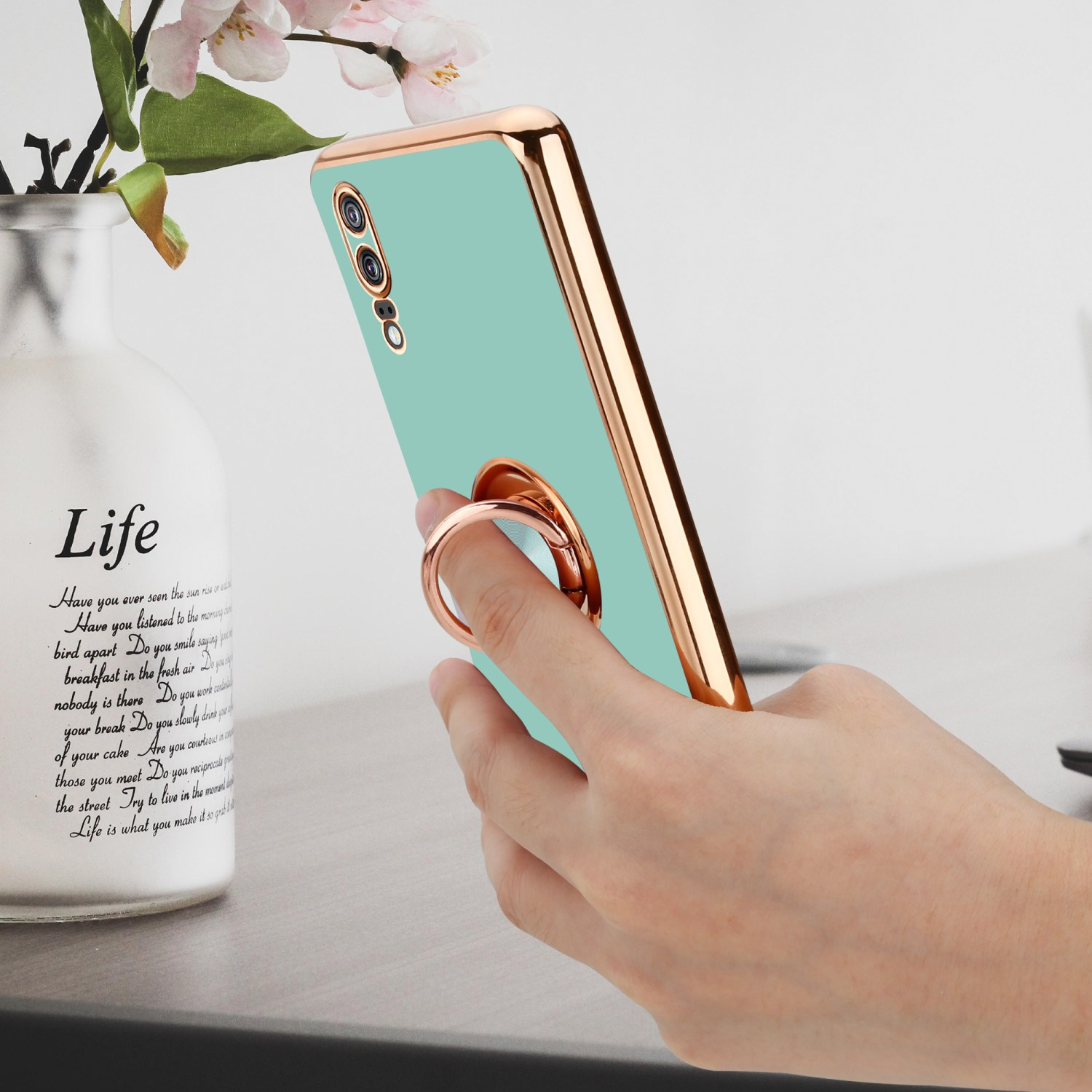 Une coque de téléphone vert menthe avec un support de bague doré à l'arrière, tenue par une main, avec un vase en arrière-plan.