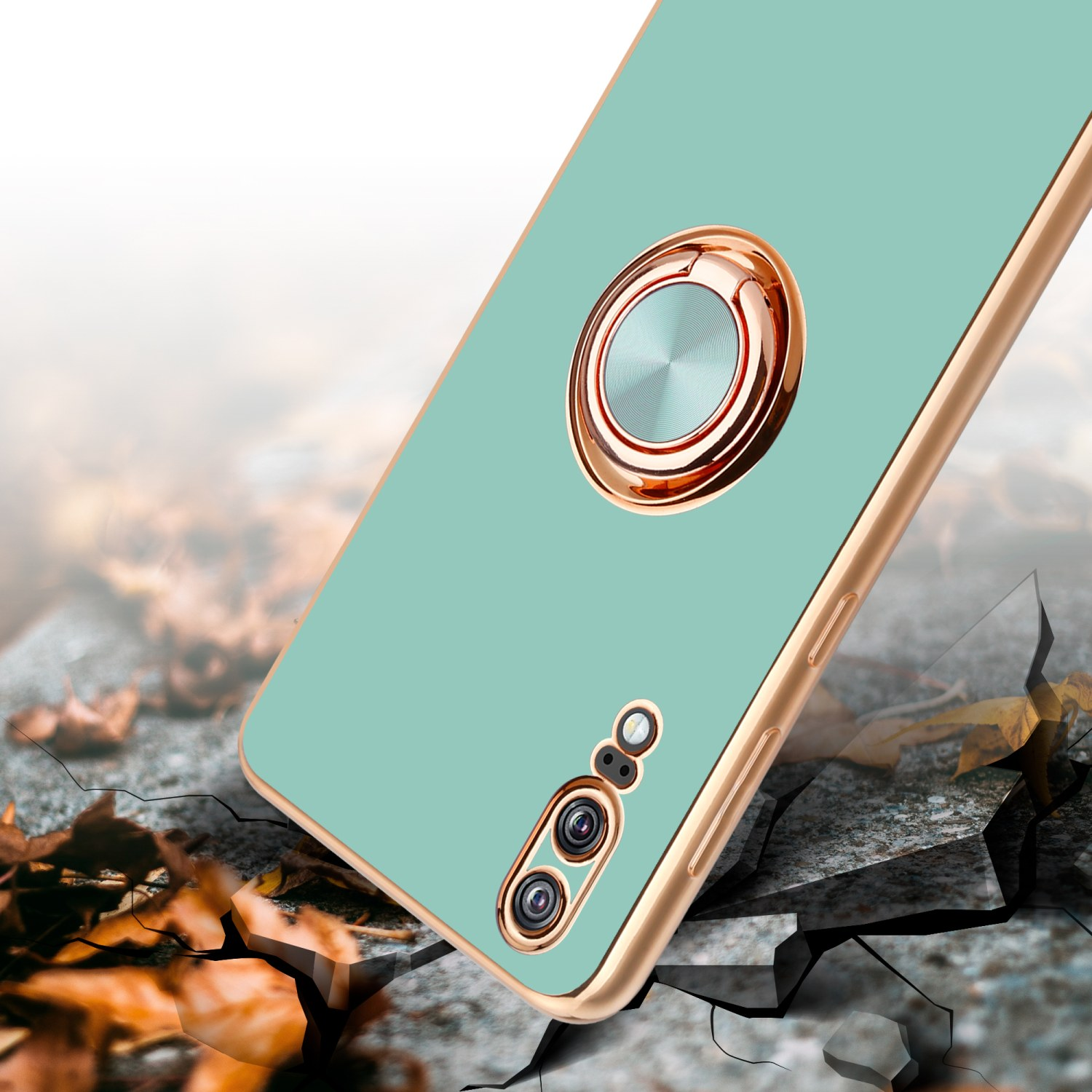 Une coque de téléphone vert menthe avec un cadre doré et un support d'anneau repose sur un sol fissuré. Il y a des feuilles autour.