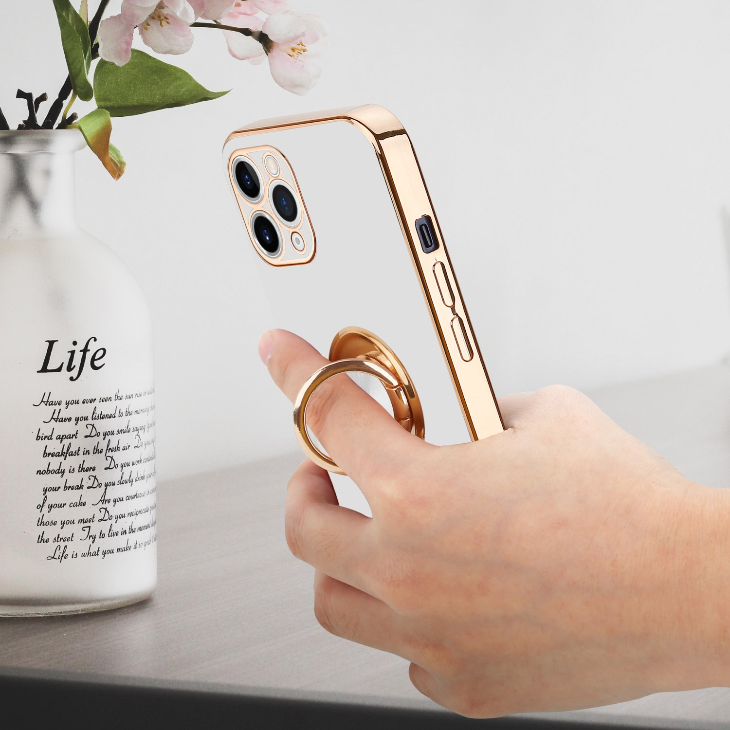 Eine Hand hält eine goldene Handyhülle mit Ring, neben einer Vase mit Blumen und Text auf einem Tisch.