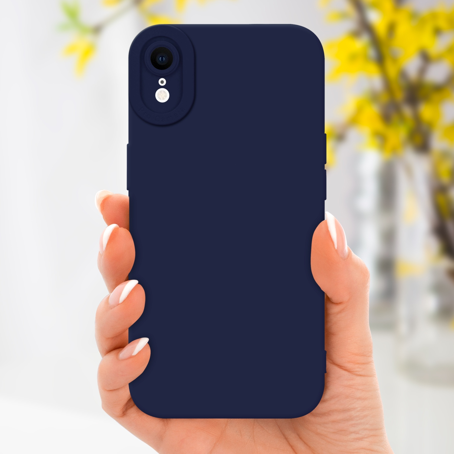 La mano di una donna che tiene una custodia blu navy con un ritaglio per la fotocamera, davanti a fiori gialli sfocati.