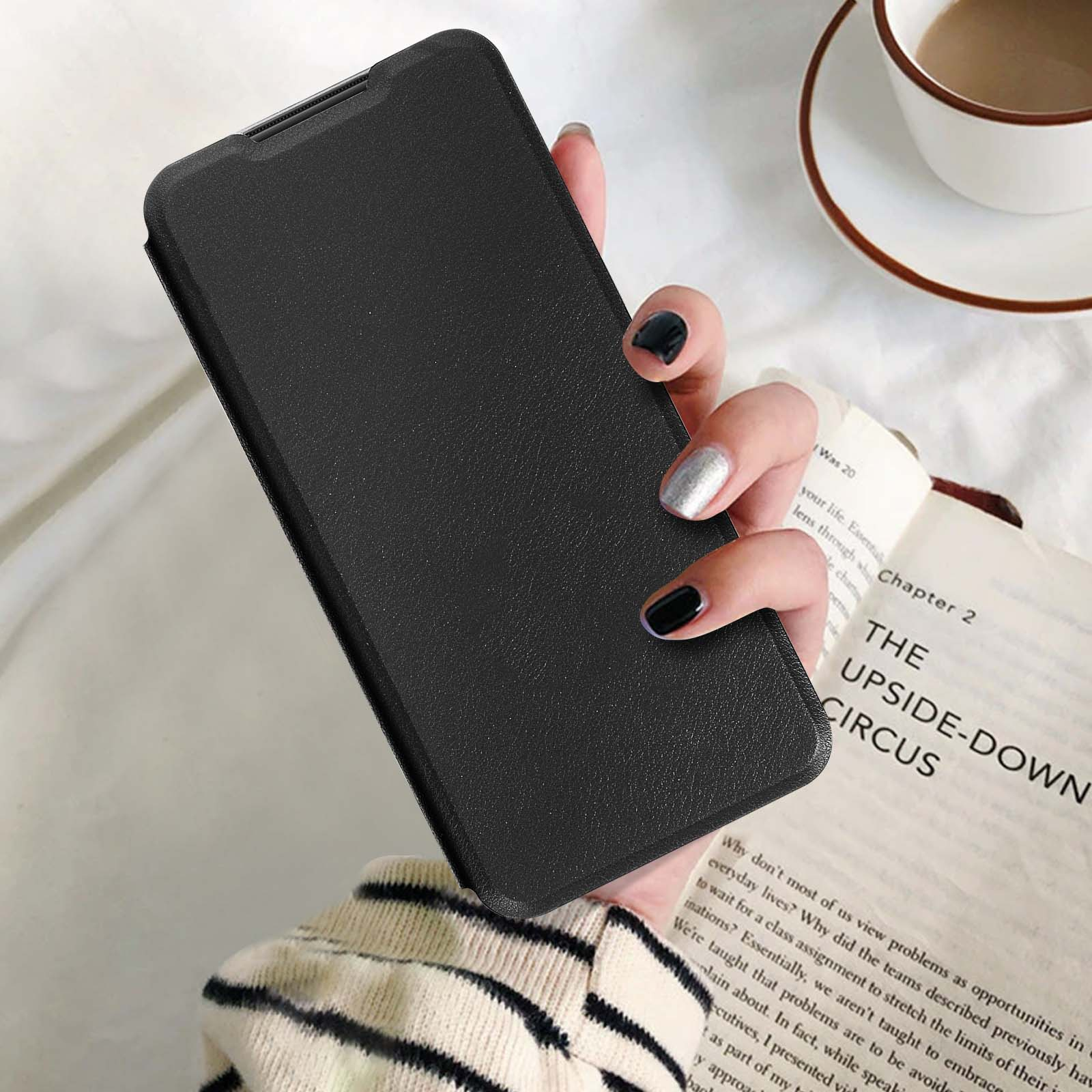 Hand hält eine geschlossene schwarze Handyhülle. Offenes Buch und Kaffeetasse im Hintergrund.