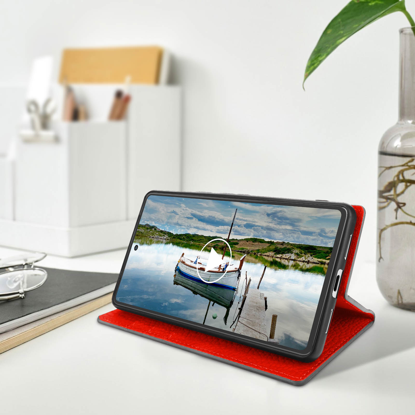 Telefon in roter Hülle mit See-Szene mit Boot. Weißer Tisch, Vase und Objekte im Hintergrund.