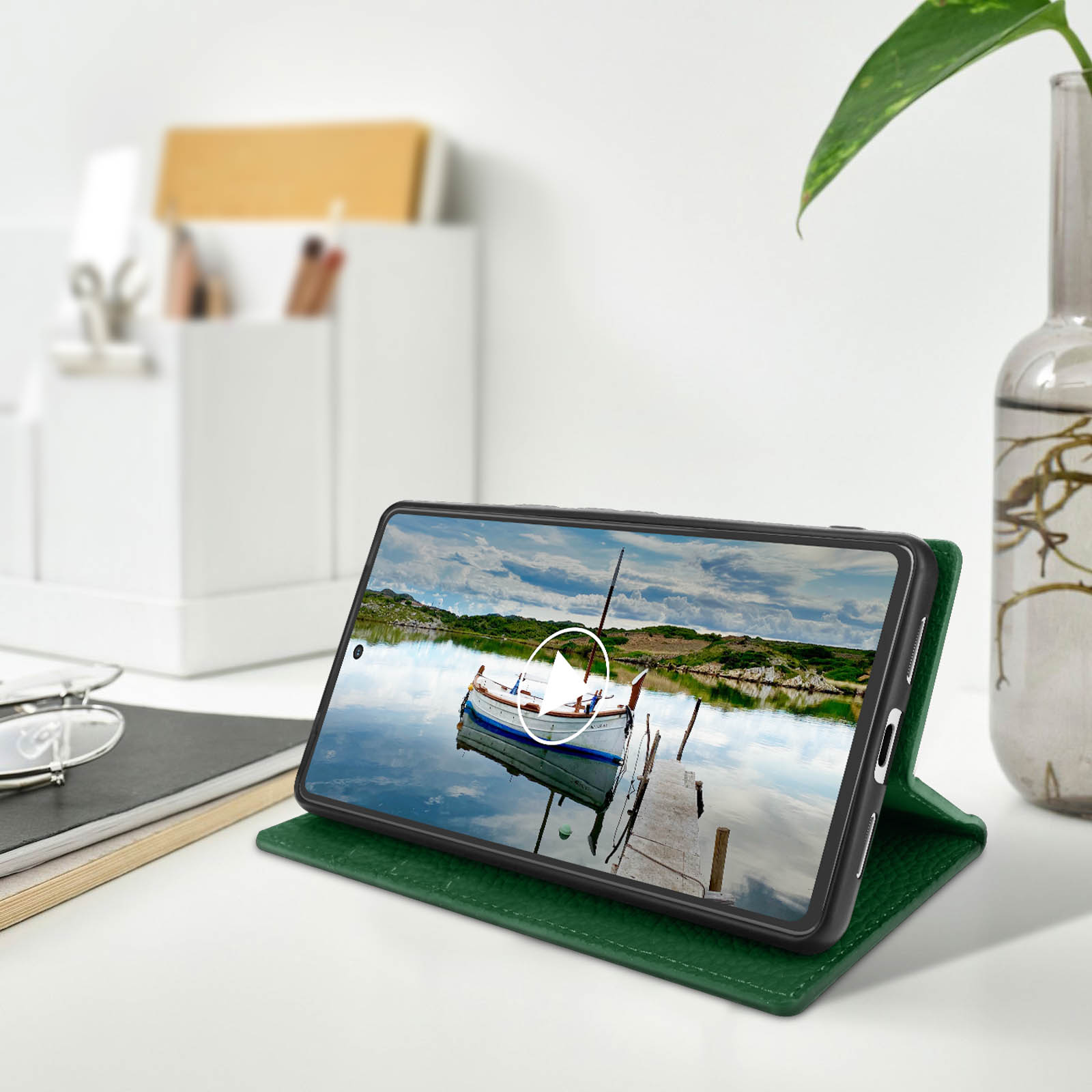 Un téléphone dans un étui vert est sur une table, affichant une scène de lac et de bateau. Un vase et des lunettes sont à proximité.