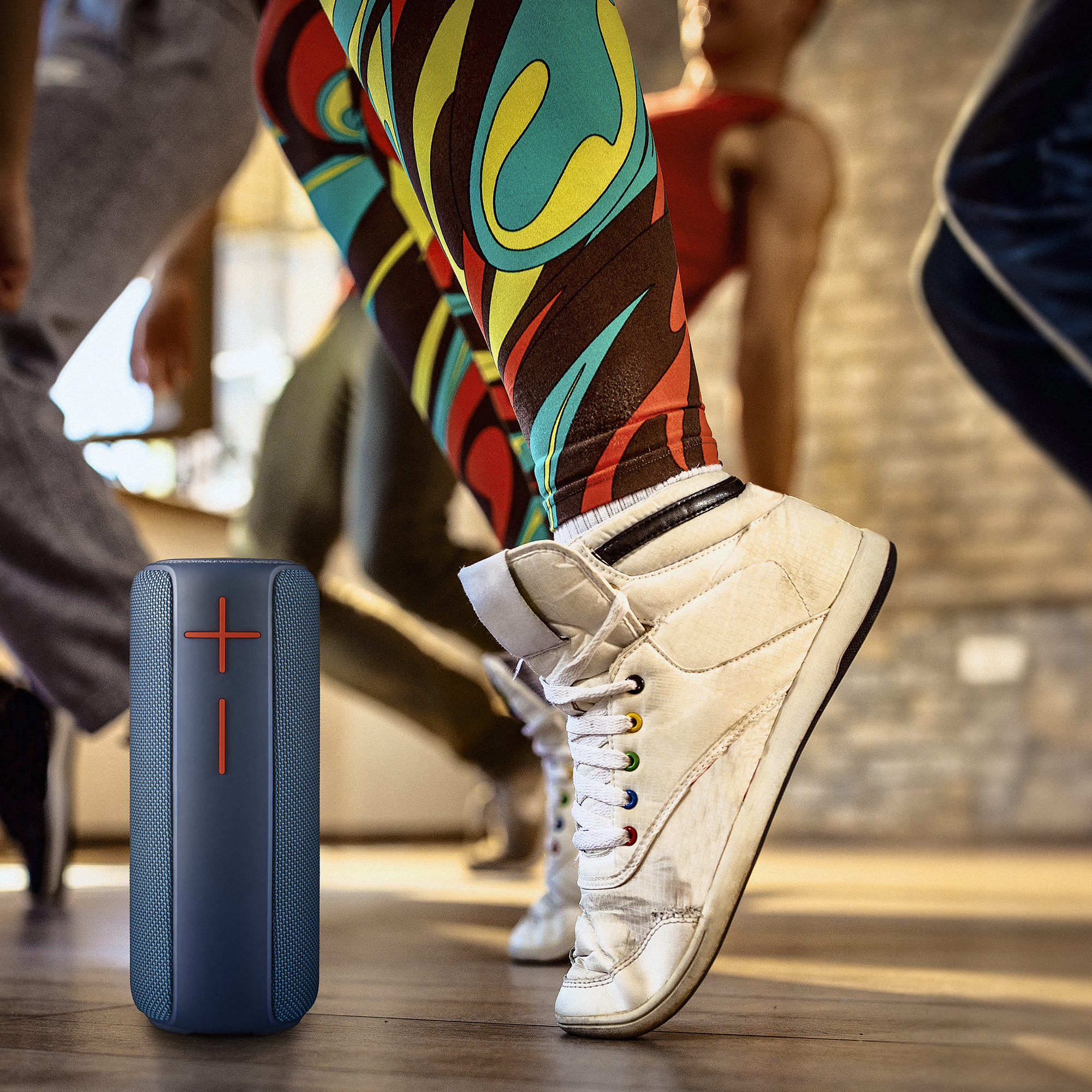 Altavoz portátil azul con detalles naranjas, pies de bailarines al fondo.
