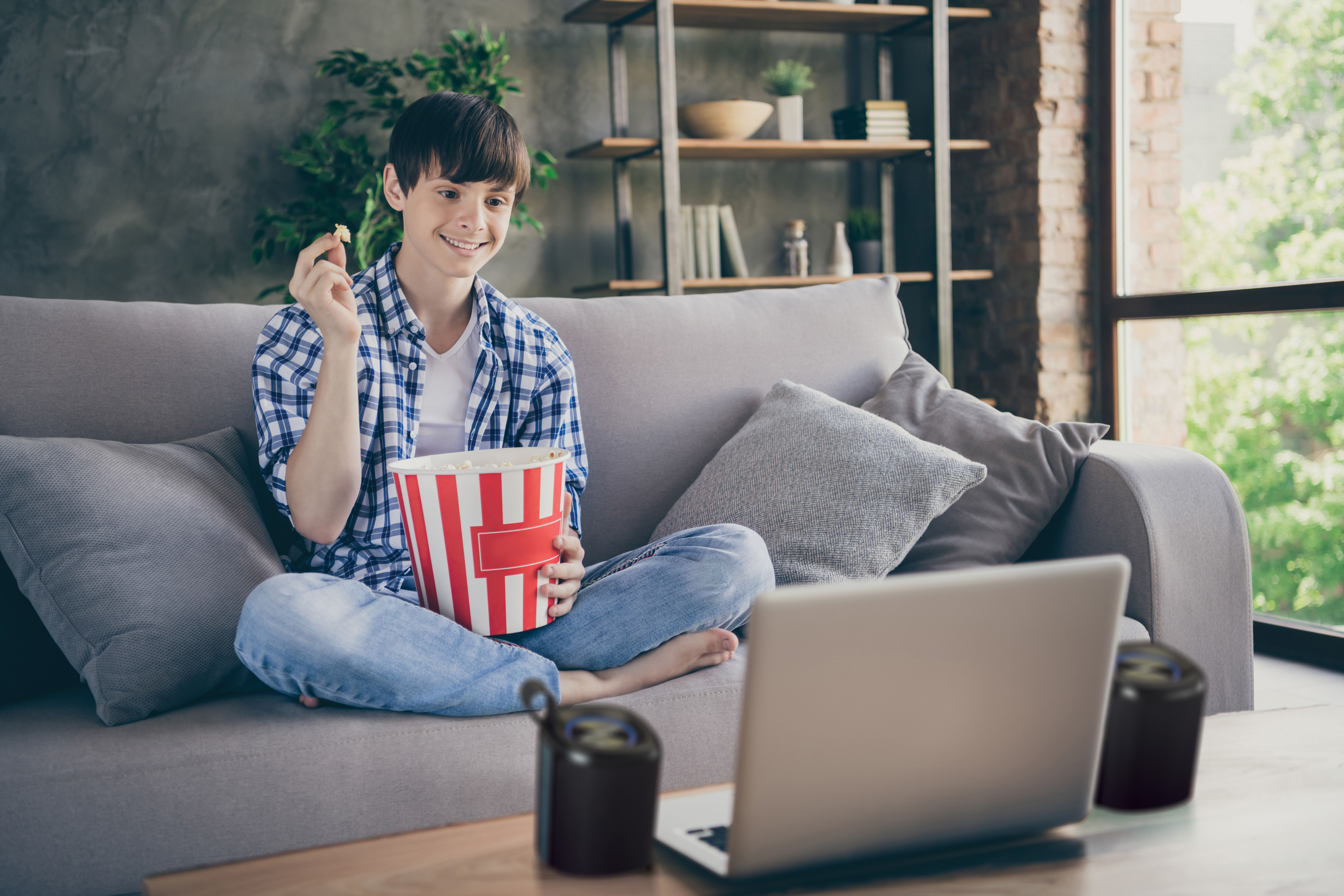 Mężczyzna siedzi na kanapie, ogląda laptopa, je popcorn.