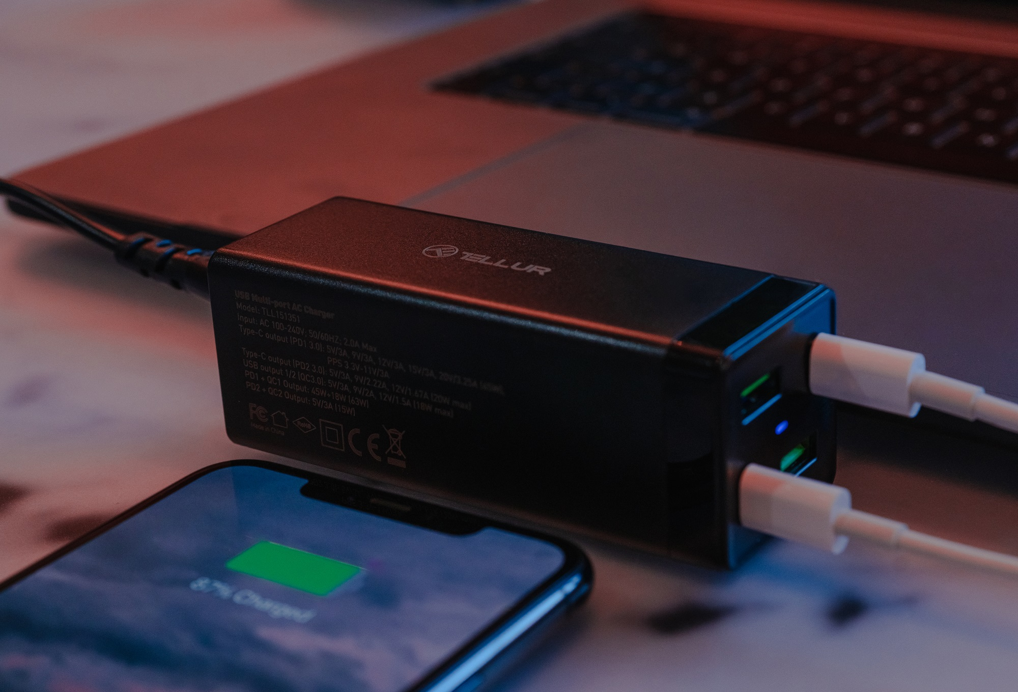 Czarna ładowarka z wieloma portami, ładująca telefon i laptop w tle.