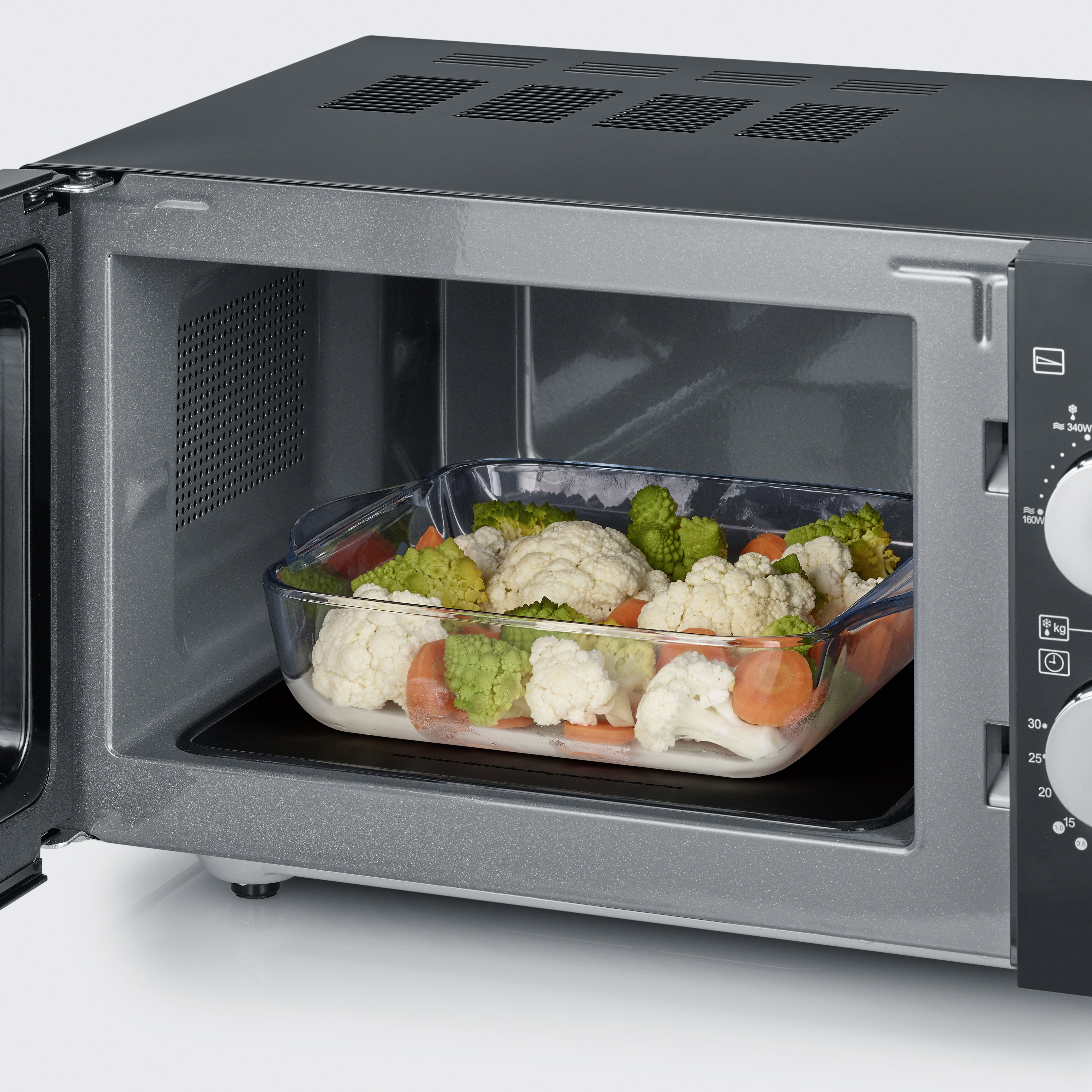 Kuchenka mikrofalowa z otwartymi drzwiami. W środku szklane naczynie zawiera warzywa.