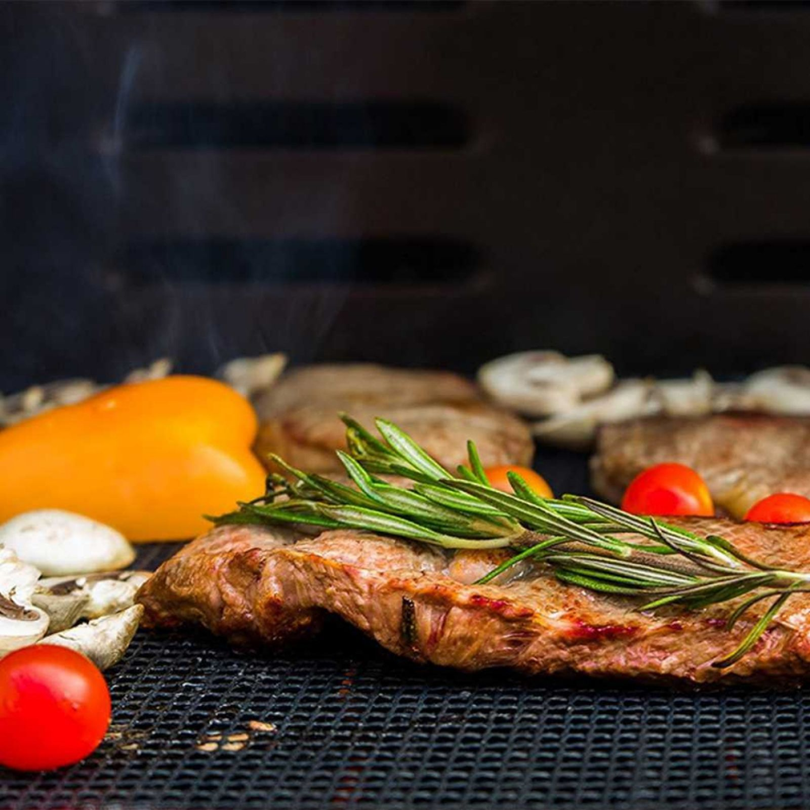 Gegrilltes Steak mit Rosmarin, Tomaten und gelber Paprika auf einem Grill. Rauch steigt auf.