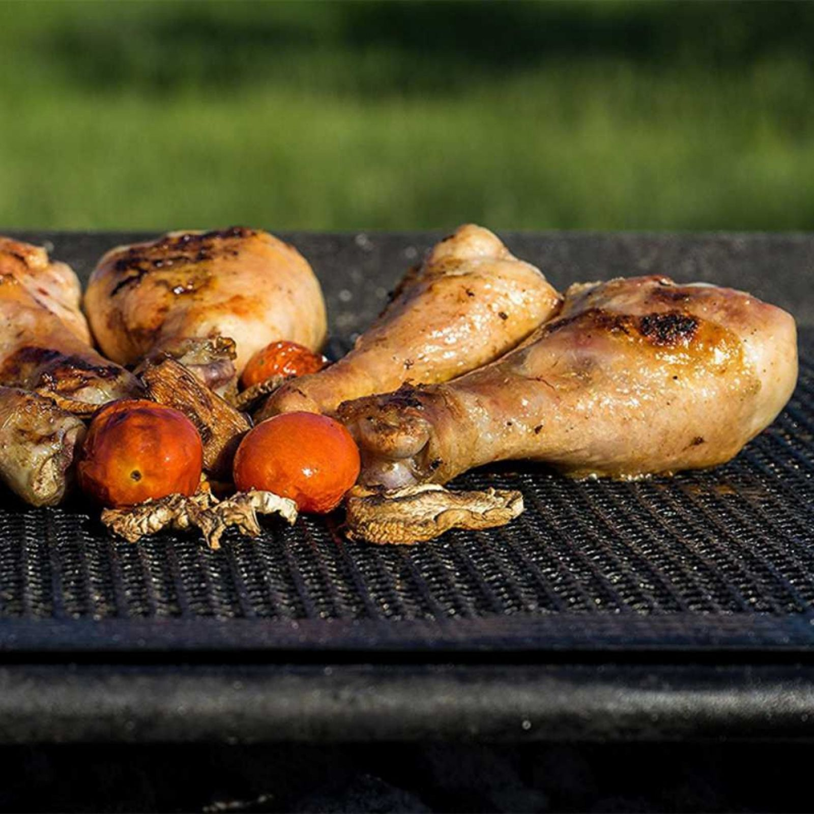 Gegrillte Hähnchenschenkel, Tomaten und Pilze auf einem Grill. Grünes Gras im Hintergrund sichtbar.