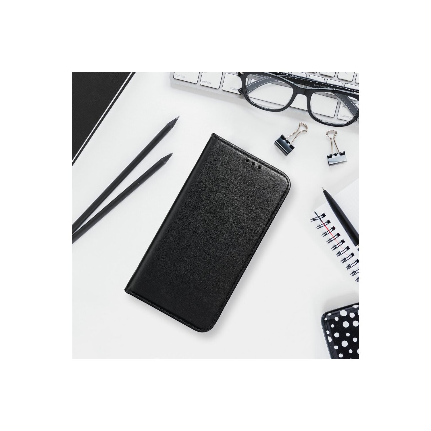 Eine schwarze Handyhülle, Bleistifte, Brille, Stift und Notizbuch liegen auf weißem Untergrund.