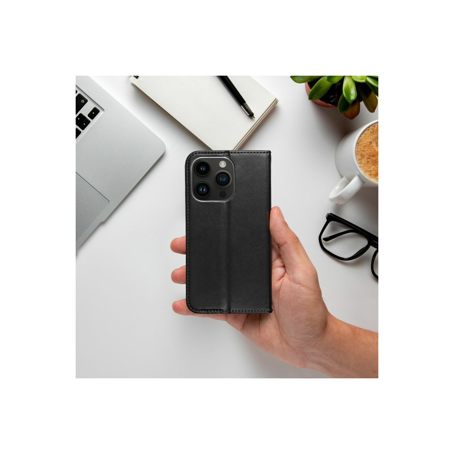 Eine Hand, die eine schwarze Handyhülle mit einem Telefon darin hält. Laptop, Brille und eine Kaffeetasse sind sichtbar.