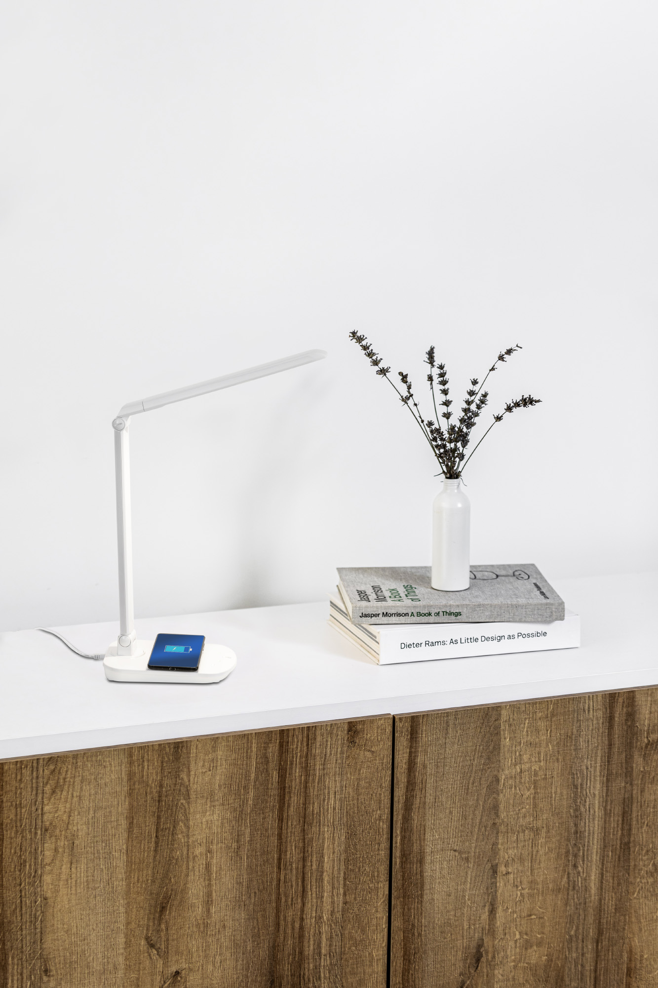 Una lampada da scrivania bianca, un telefono in carica e libri con un vaso di fiori.
