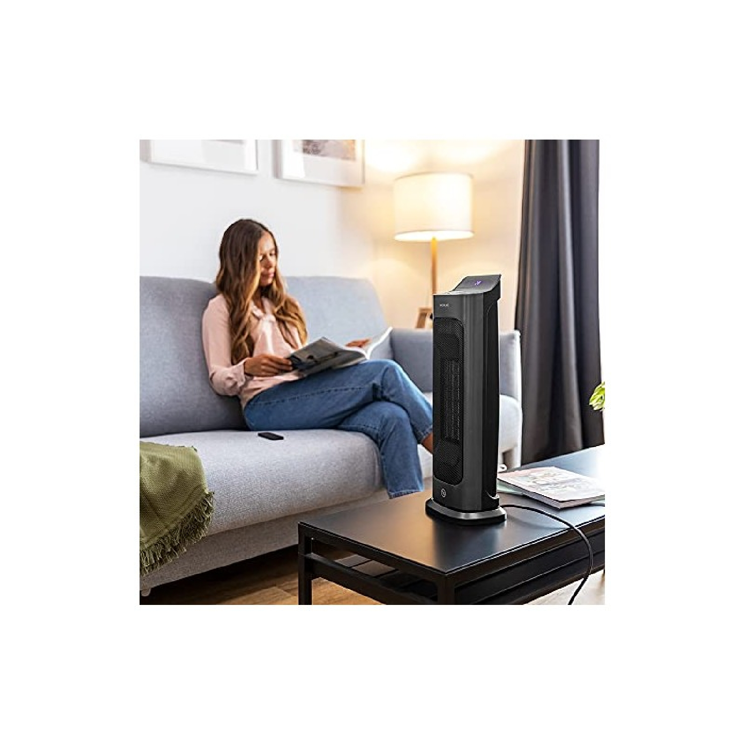 Kobieta siedzi na szarej sofie, czytając książkę. Czarny grzejnik stoi na stole. Lampa jest za nią.