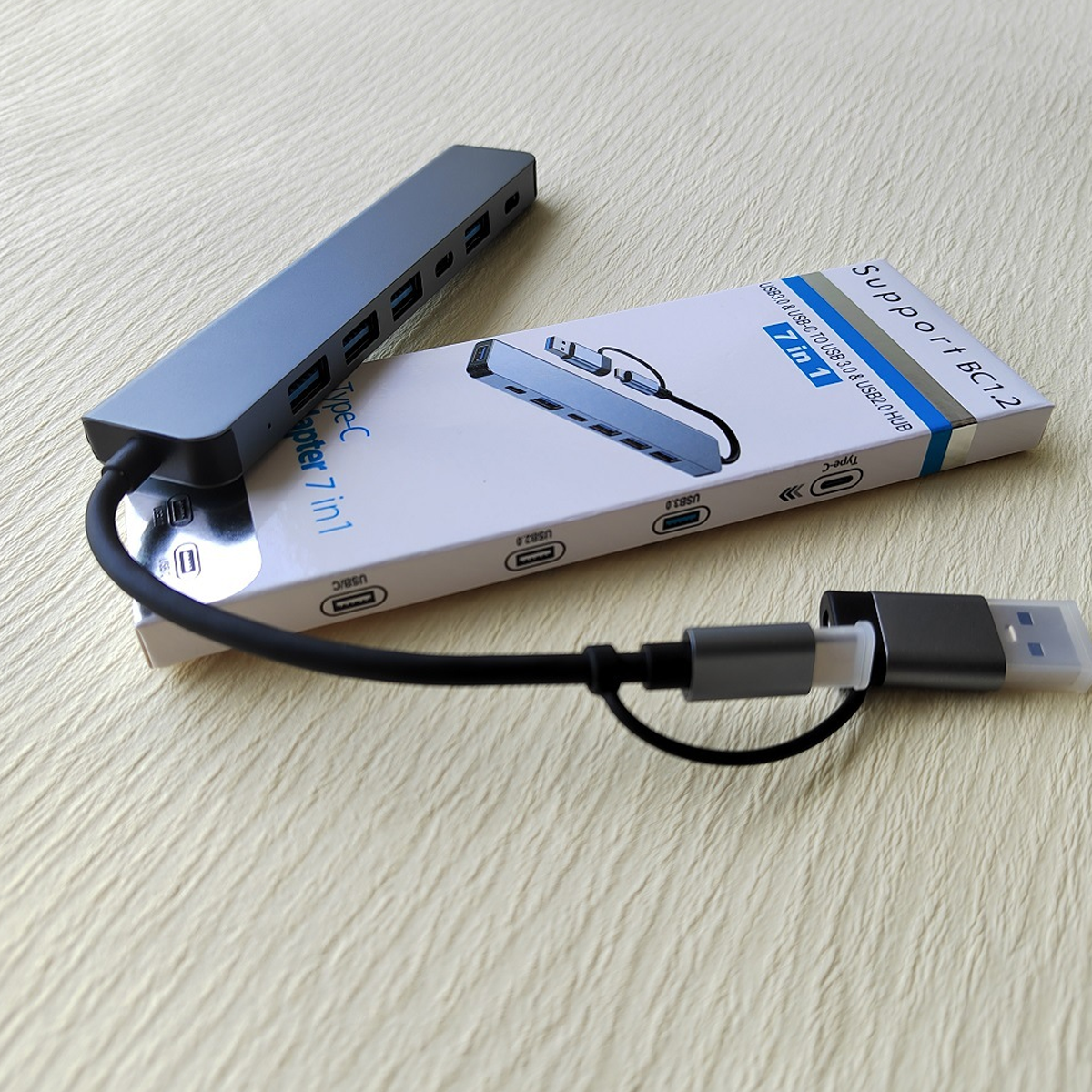 Ein grauer USB-C-Hub mit schwarzem Kabel und einer weißen Box auf einer beigefarbenen Oberfläche.